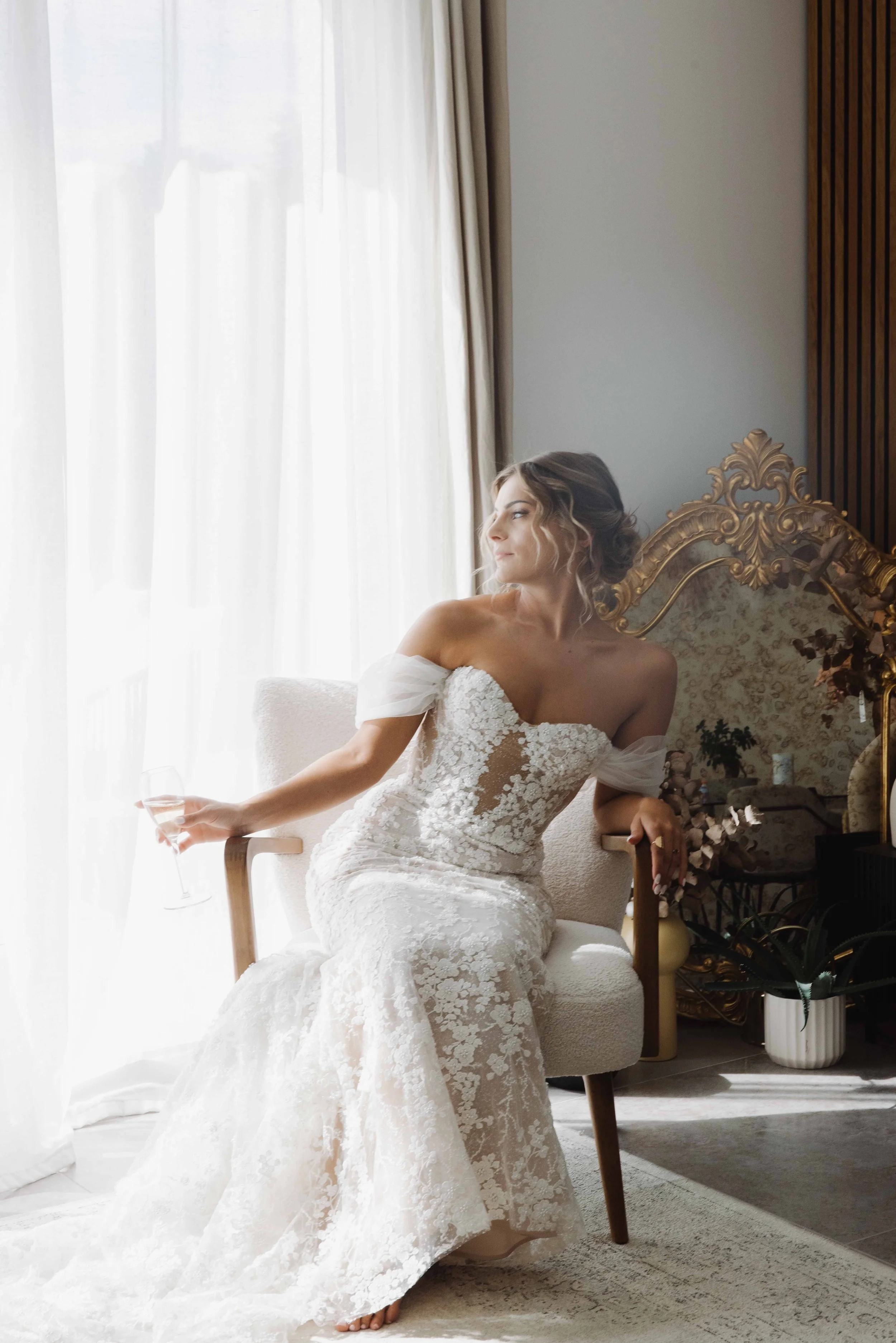 Une femme en robe de mariée blanche assise sur une chaise près d'une fenêtre, tenant un verre de champagne, dans une pièce élégante avec des draps blancs et un décor luxueux. Photographe mariage Isère, ferme du rocher, cérémonie civil et laïque. Lyon