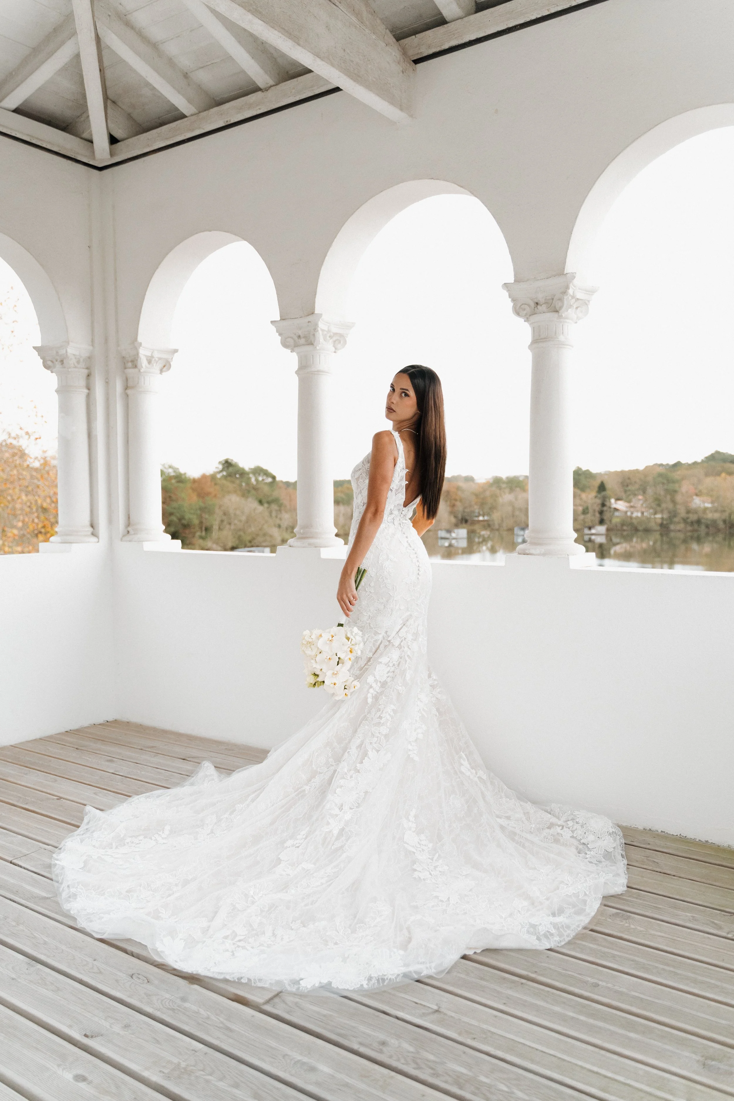 Une femme en robe de mariée blanche tenant un bouquet de fleurs, debout sur une terrasse avec des arches blanches et un paysage de lac et de arbres en arrière-plan.