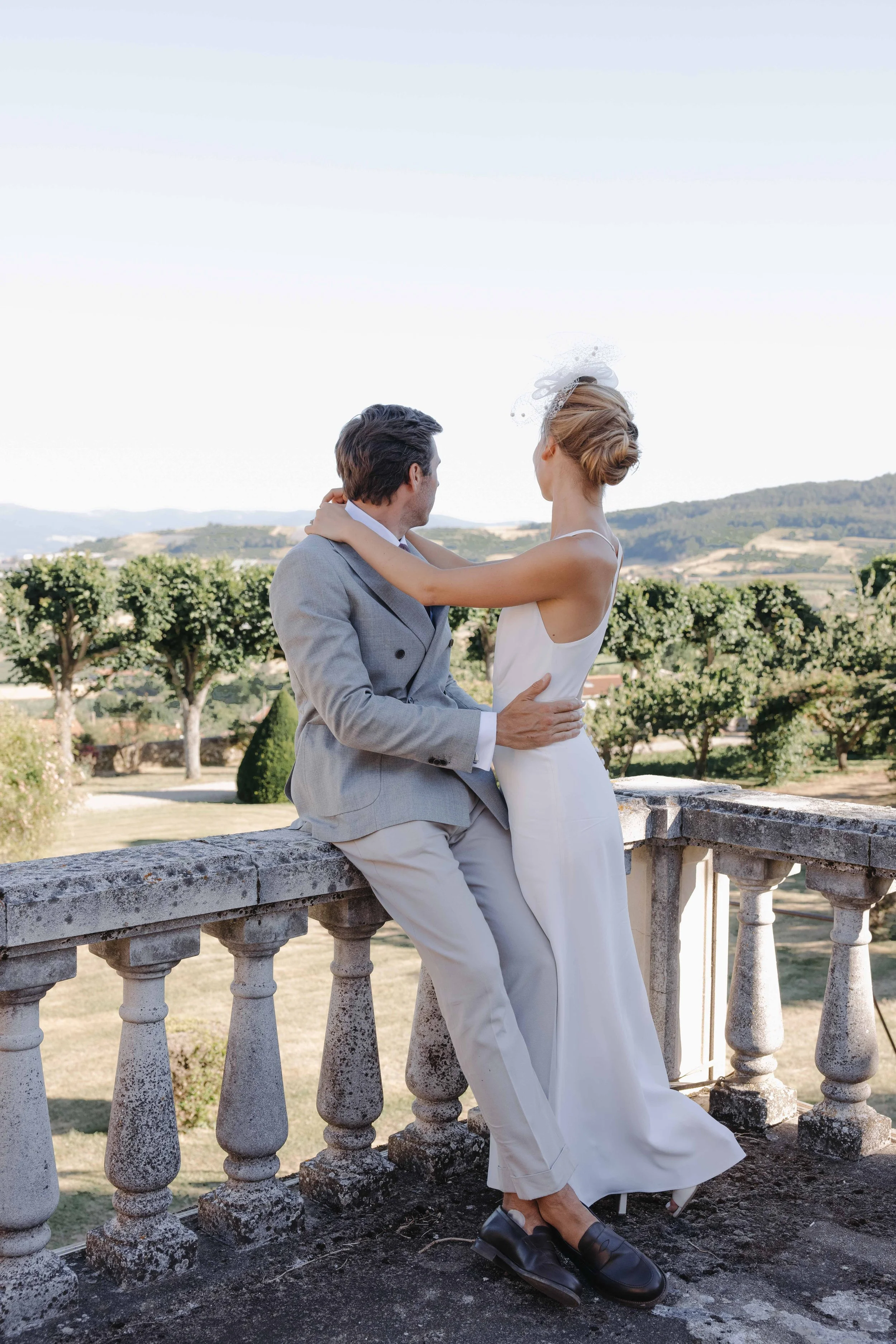 Un couple en tenue de mariage assis sur une balustrade en pierre, regardant l'un l'autre, avec un paysage de campagne en arrière-plan dans le beaujolais à côté de Lyon, rhone alpes, lors d'un mariage dans un chateau