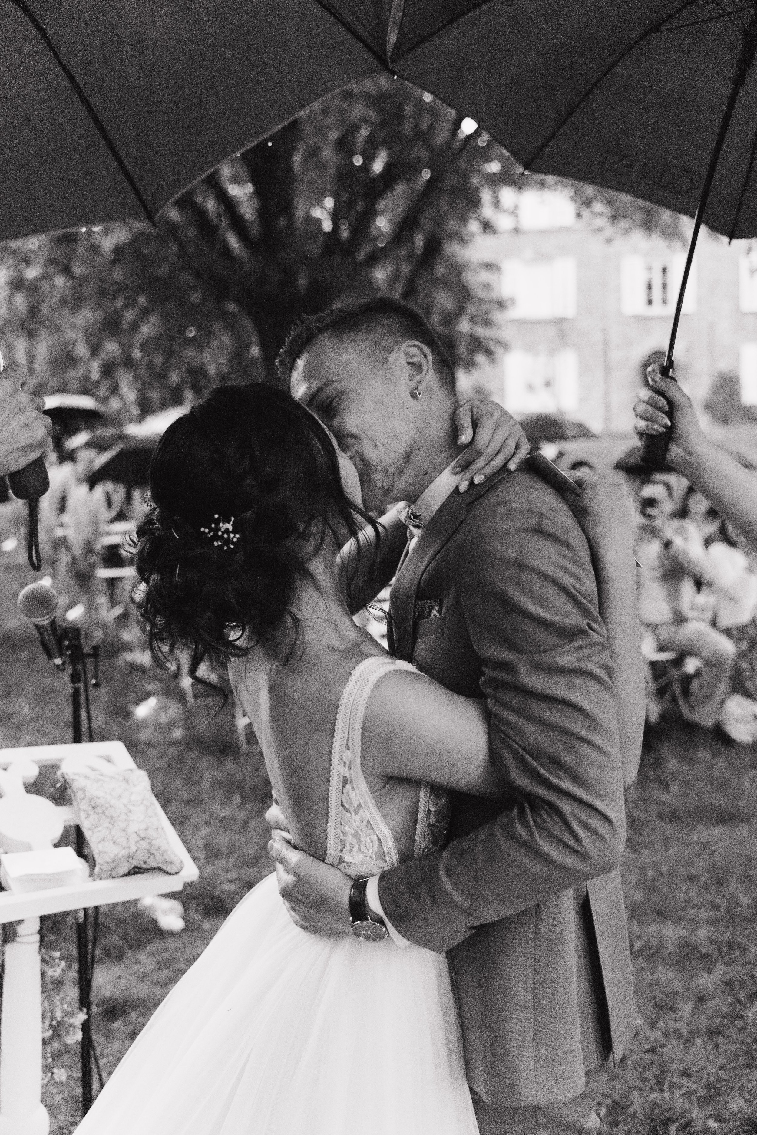 Un couple marié échangeant un baiser sous un parapluie lors d'une cérémonie en extérieur, une foule de personnes en arrière-plan. Domaine de tourieux à Savigny dans le beaujolais à Lyon. Mariage cérémonie laïque, photographe Lyon haut de gamme, duo