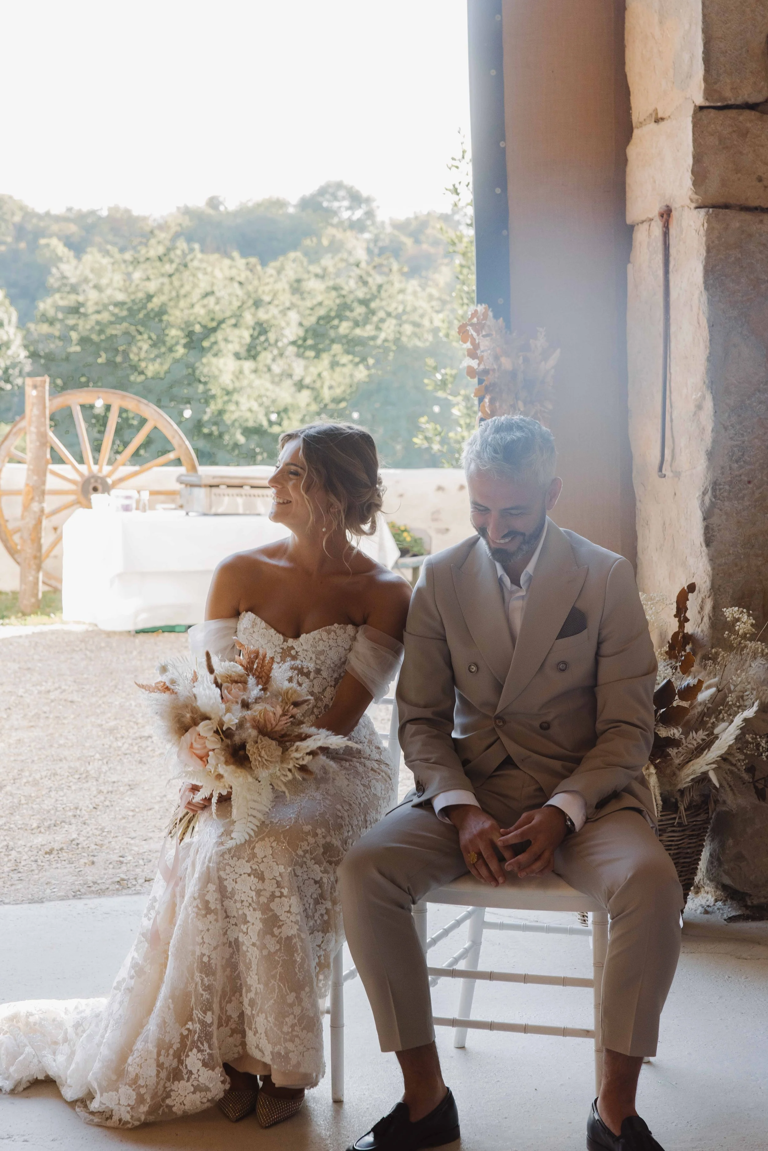 Un couple de mariés assis ensemble dans une salle lumineuse lors d'un mariage, devant un grand ouvert avec une vue de la nature en arrière-plan. Photographe mariage Isère, ferme du rocher, cérémonie civil et laïque. Bordeaux et Lyon et destination.