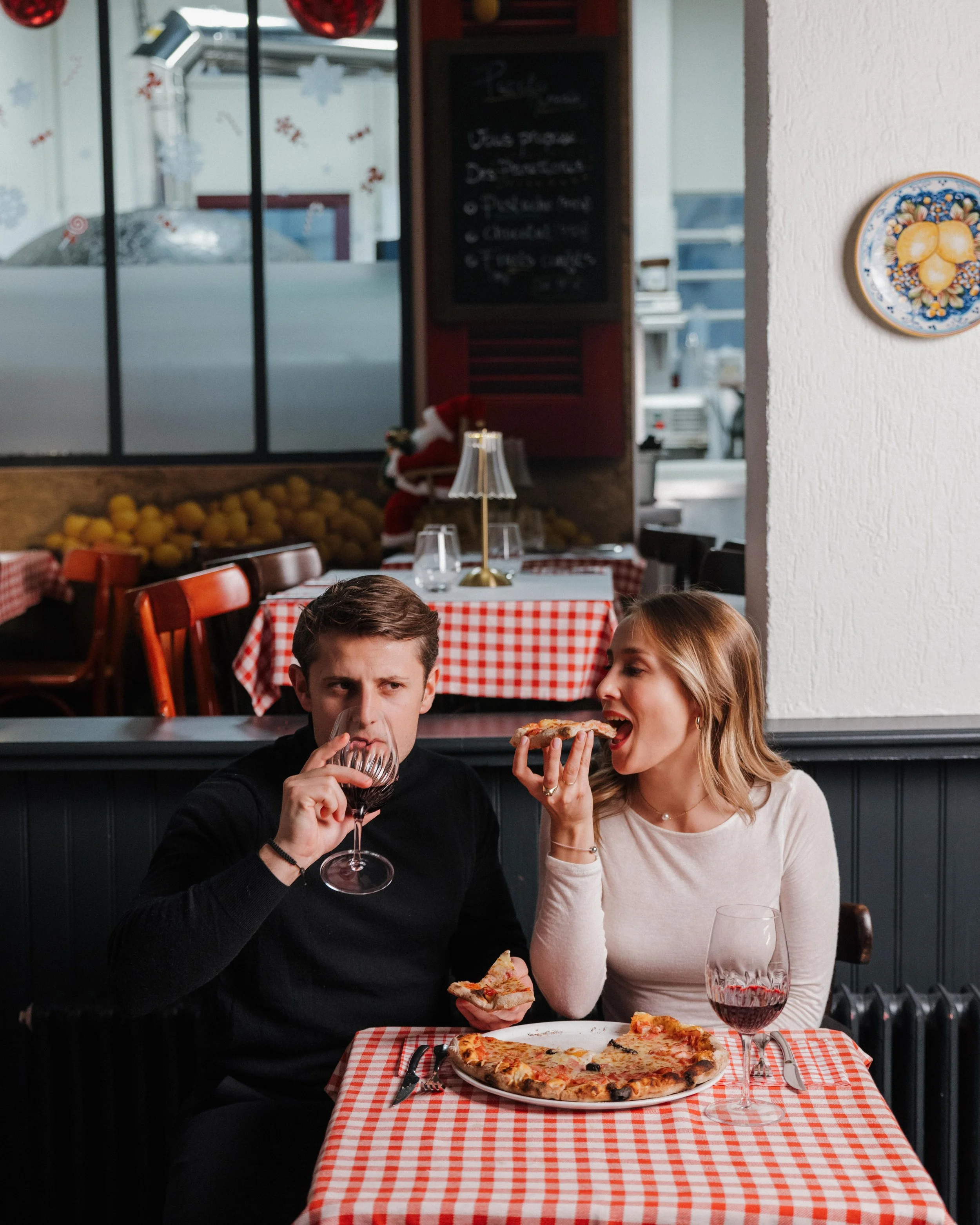 Deux personnes, un homme et une femme, dégustent de la pizza dans un restaurant italien. La femme tient une part de pizza près de sa bouche, et l'homme boit du vin rouge. La table est couverte d'une nappe à carreaux rouges et blancs, avec une pizza, 