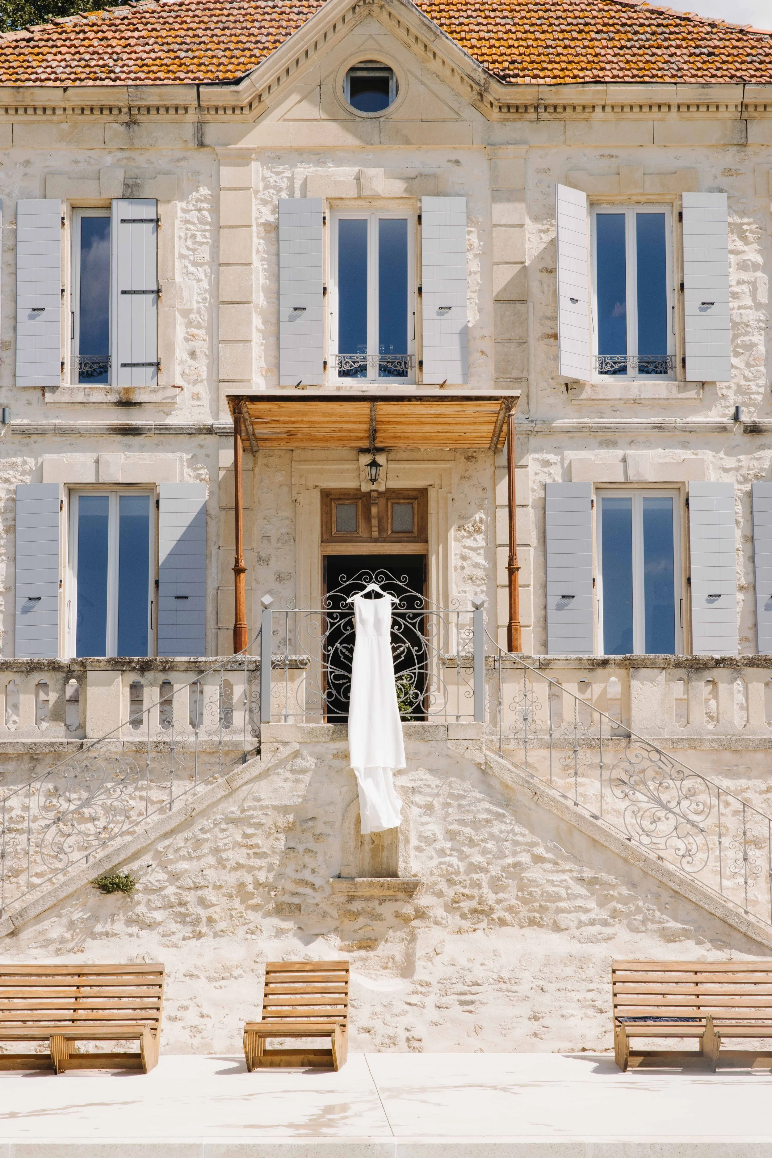 Une maison en pierre avec plusieurs fenêtres à volets blancs, une escalier menant à une porte d'entrée, sur laquelle une robe blanche est suspendue. Il y a aussi des bancs en bois devant la maison. Domaine du bijoutier à grignan, drome provençal 