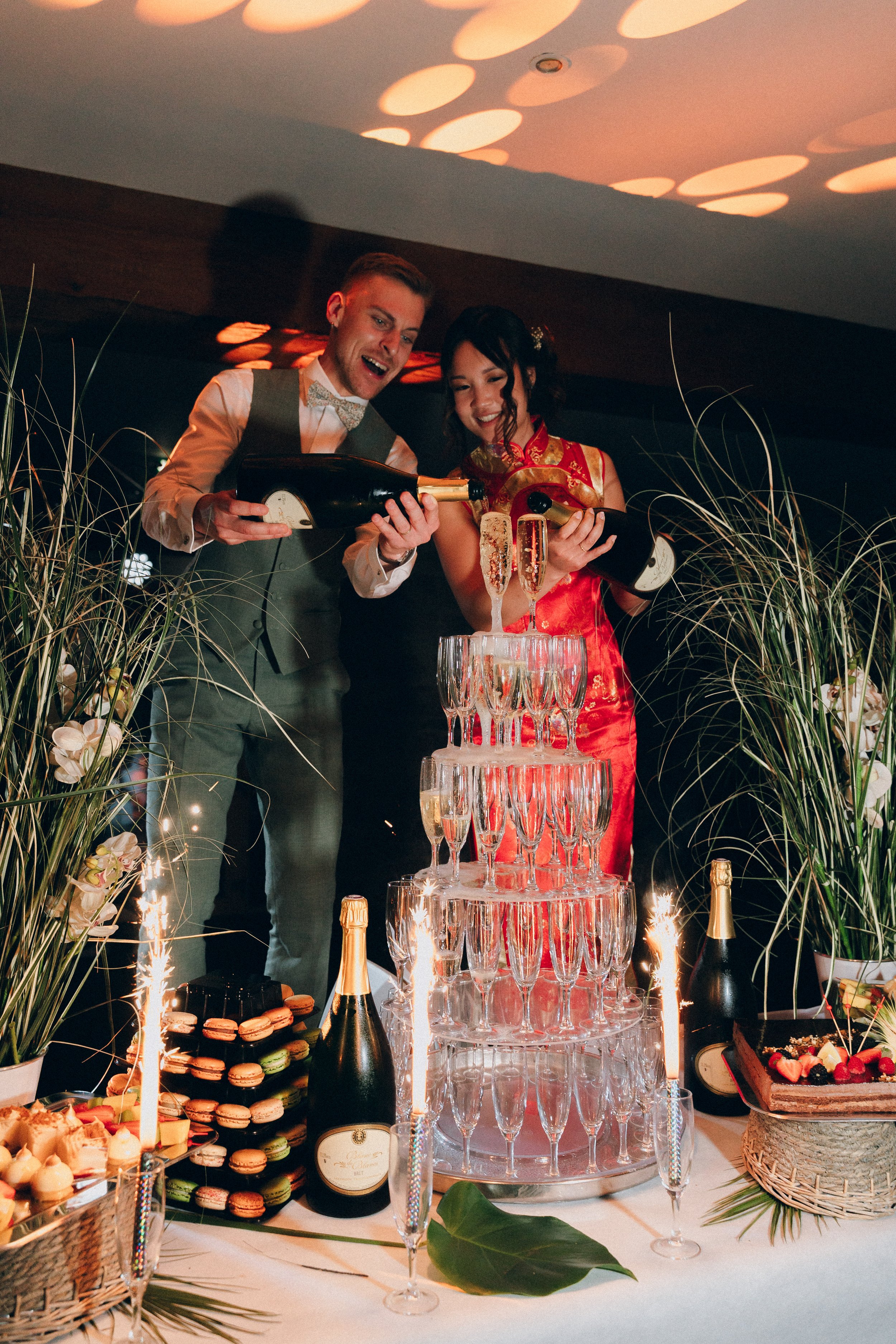 Deux personnes, un homme et une femme, célèbrent en versant du champagne dans une pyramide de verres lors d'une fête. La scène est décorée avec des plantes et des desserts. Domaine de tourieux à Savigny dans le beaujolais à Lyon. Mariage cérémonie 