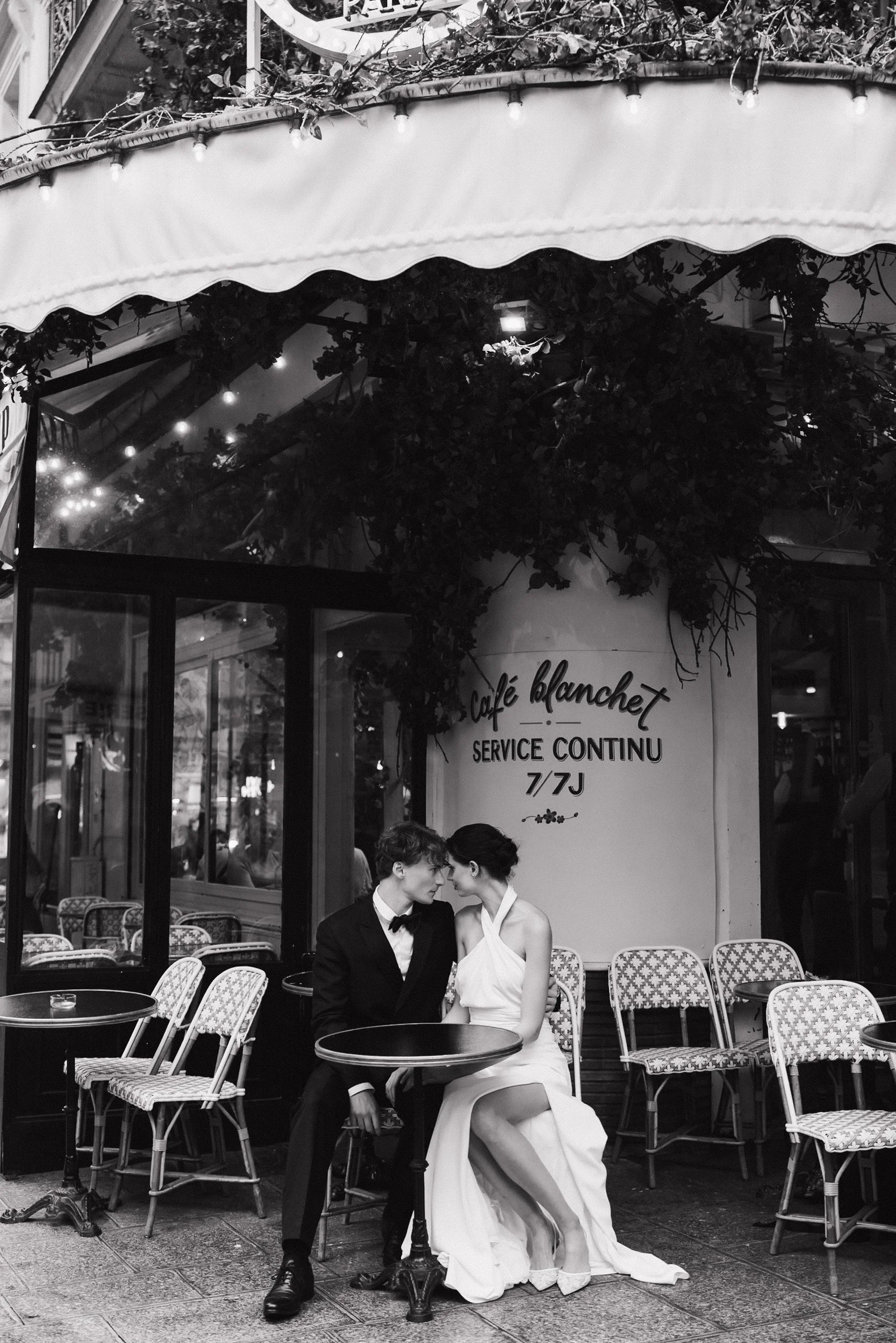 Deux personnes assises à une table devant un café parisien, vêtues élégamment, en noir et blanc. Session engagement café parisien, photographe mariage paris haut de gamme, destination wedding, madame a dit oui 
