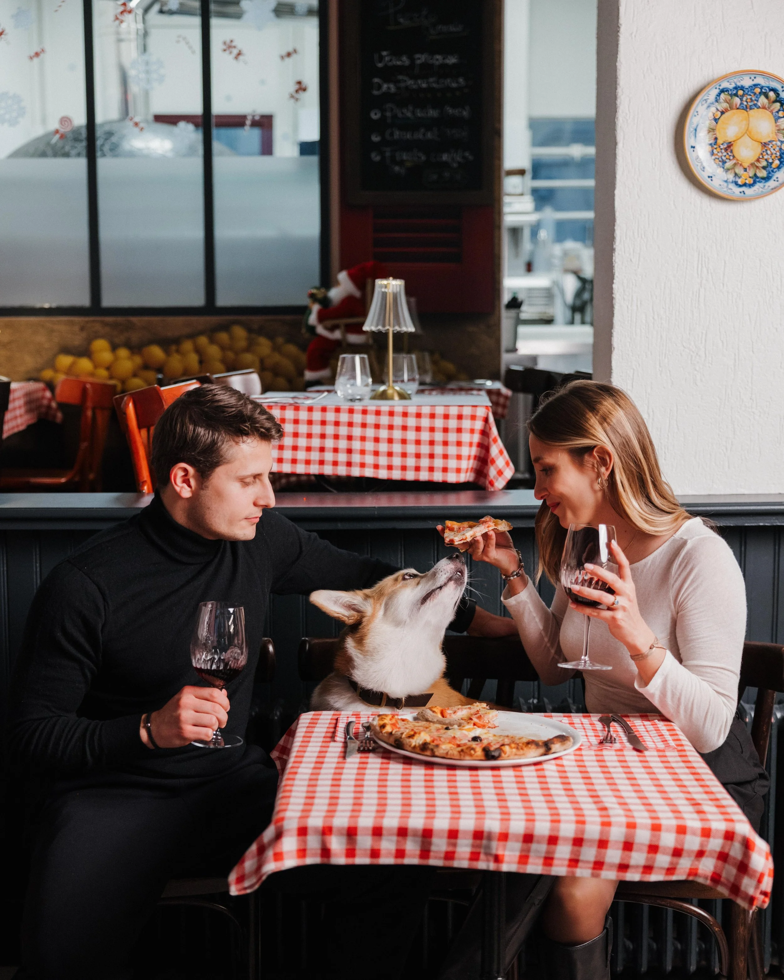 Un couple partage une pizza avec leur chien dans un restaurant, la femme lui tend une tranche de pizza. session engagement couple Lyon 6eme, brotteaux, photographe mariage bordeaux et Lyon et destination wedding