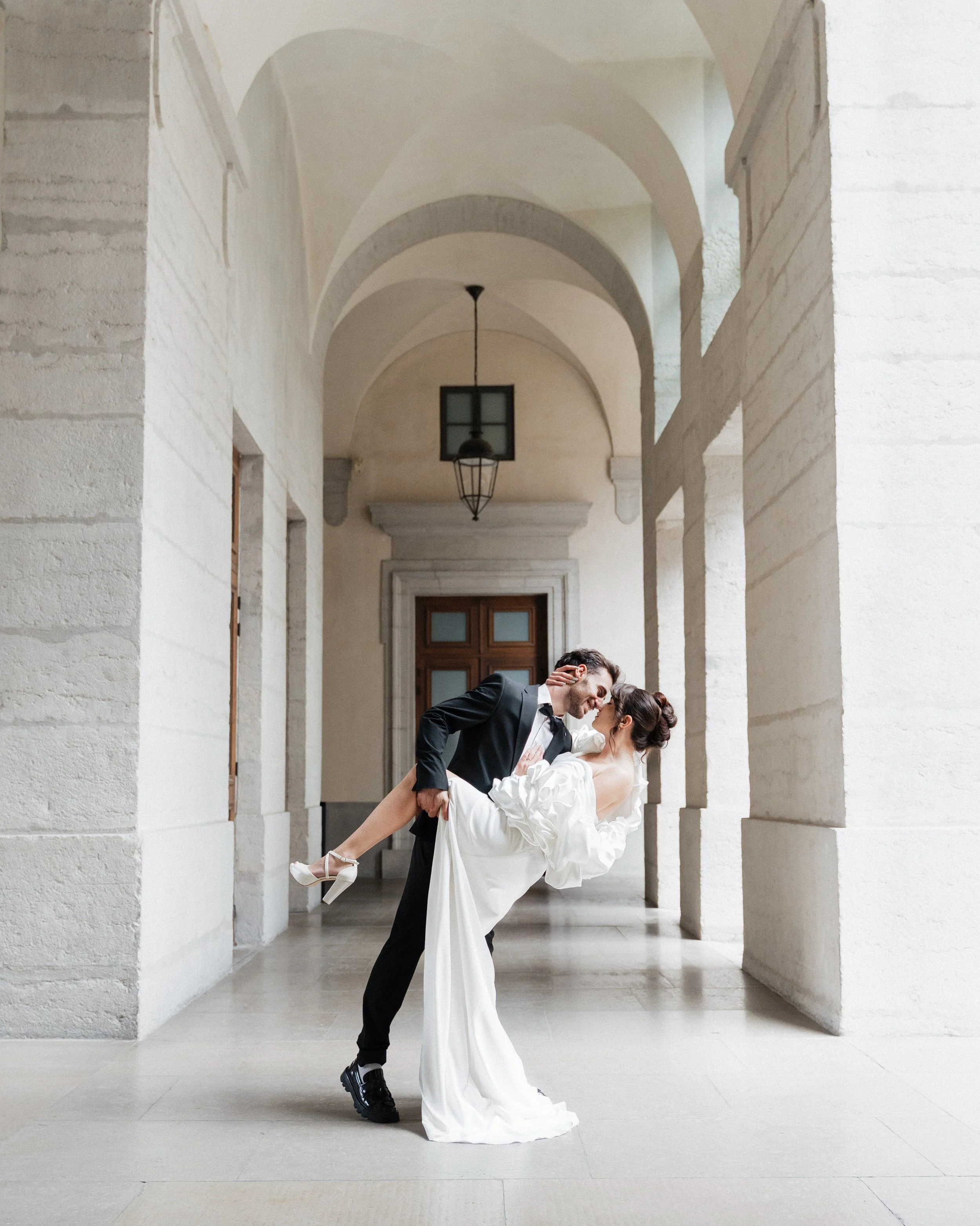 Un couple en vêtements de mariage, le homme en costume noir et la femme en robe blanche, danse dans une galerie aux arches en pierre, ambiance lumineuse et élégante à Lyon 2ème à l'hôtel dieu à l'intercontinental lors d'une session engagement.