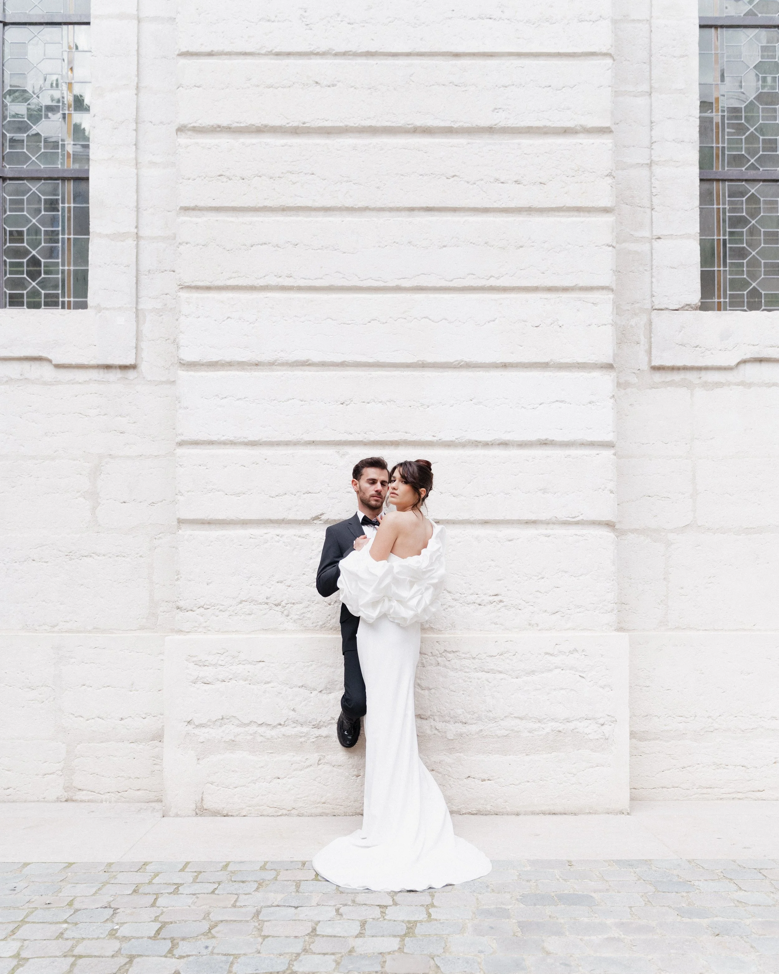 Un couple de mariés posant devant un mur blanc en pierre. La mariée porte une robe blanche longue avec des manches bouffantes, et le marié un costume sombre avec une cravate. Ils sont proches, la mariée regarde vers l'objectif, le marié regarde vers 