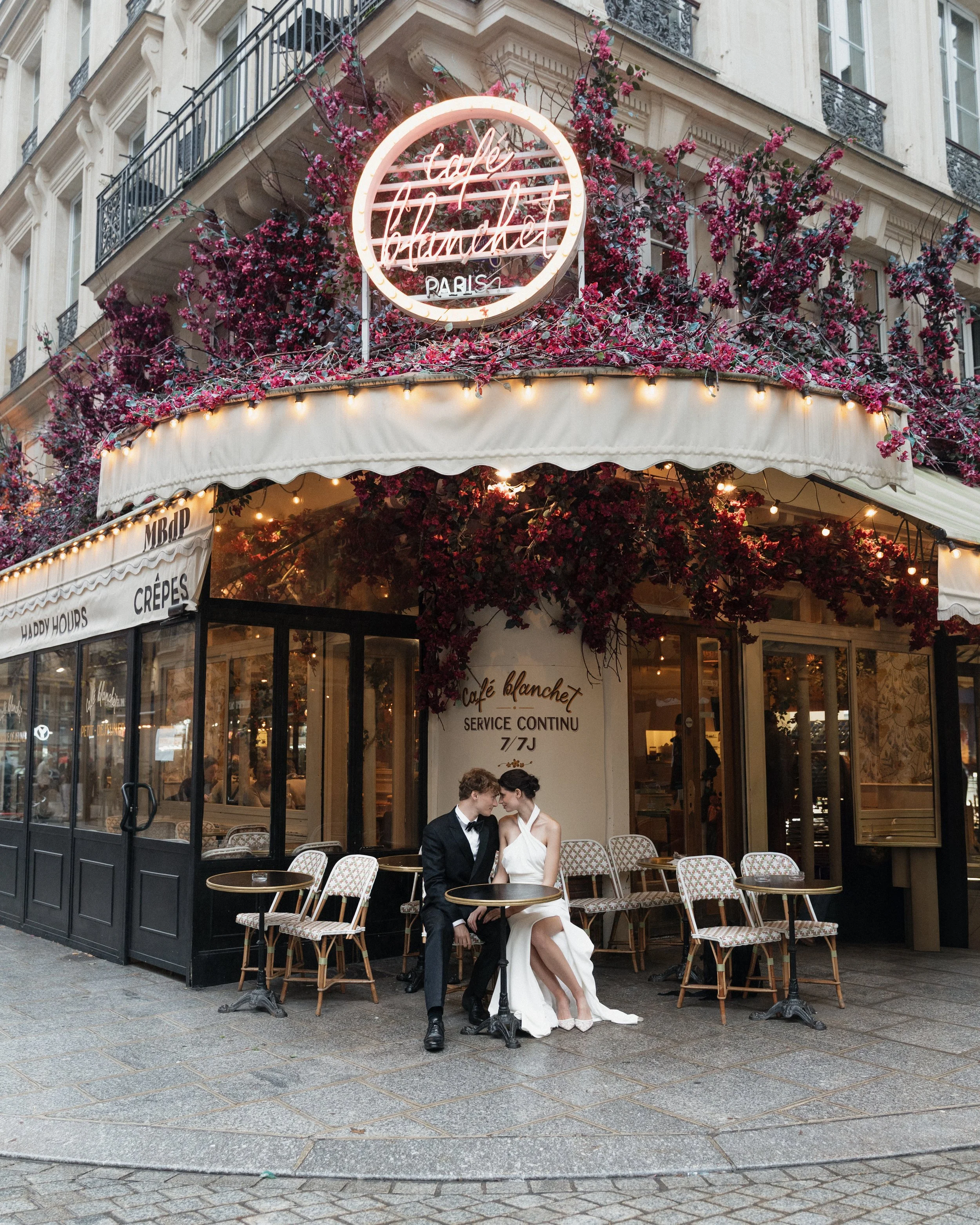 Un couple habillé élégamment, un homme en costume noir et une femme en robe blanche, assis à une table devant un café parisien appelé Café Blanchet, décoré avec des fleurs roses et des lumières, dans un style romantique. Session engagement café paris