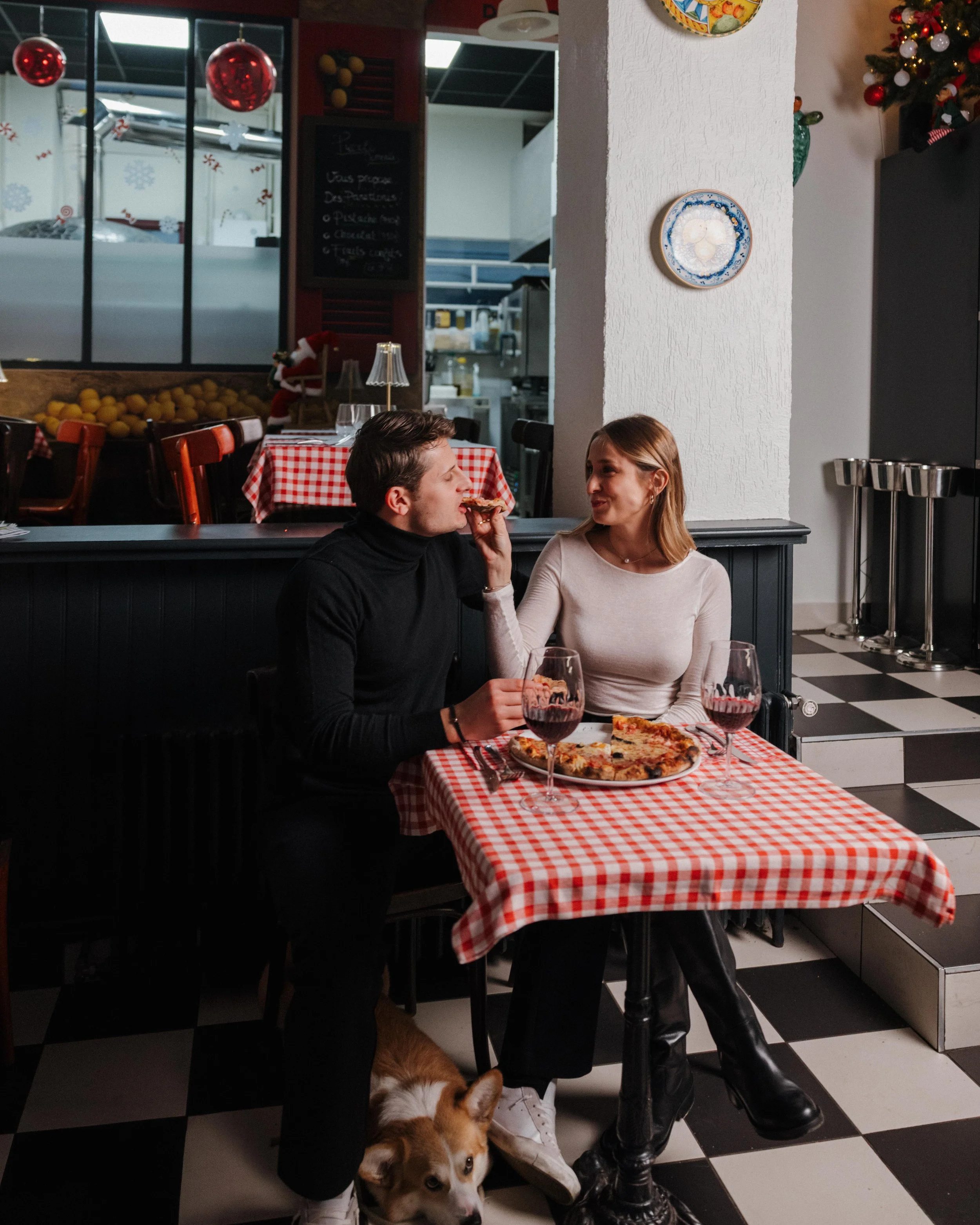 Un couple partageant une pizza dans un restaurant décoré pour Noël, avec un chien à leurs pieds et des verres de vin rouge sur la table. session engagement couple Lyon 6eme, brotteaux, photographe mariage bordeaux et Lyon et destination wedding