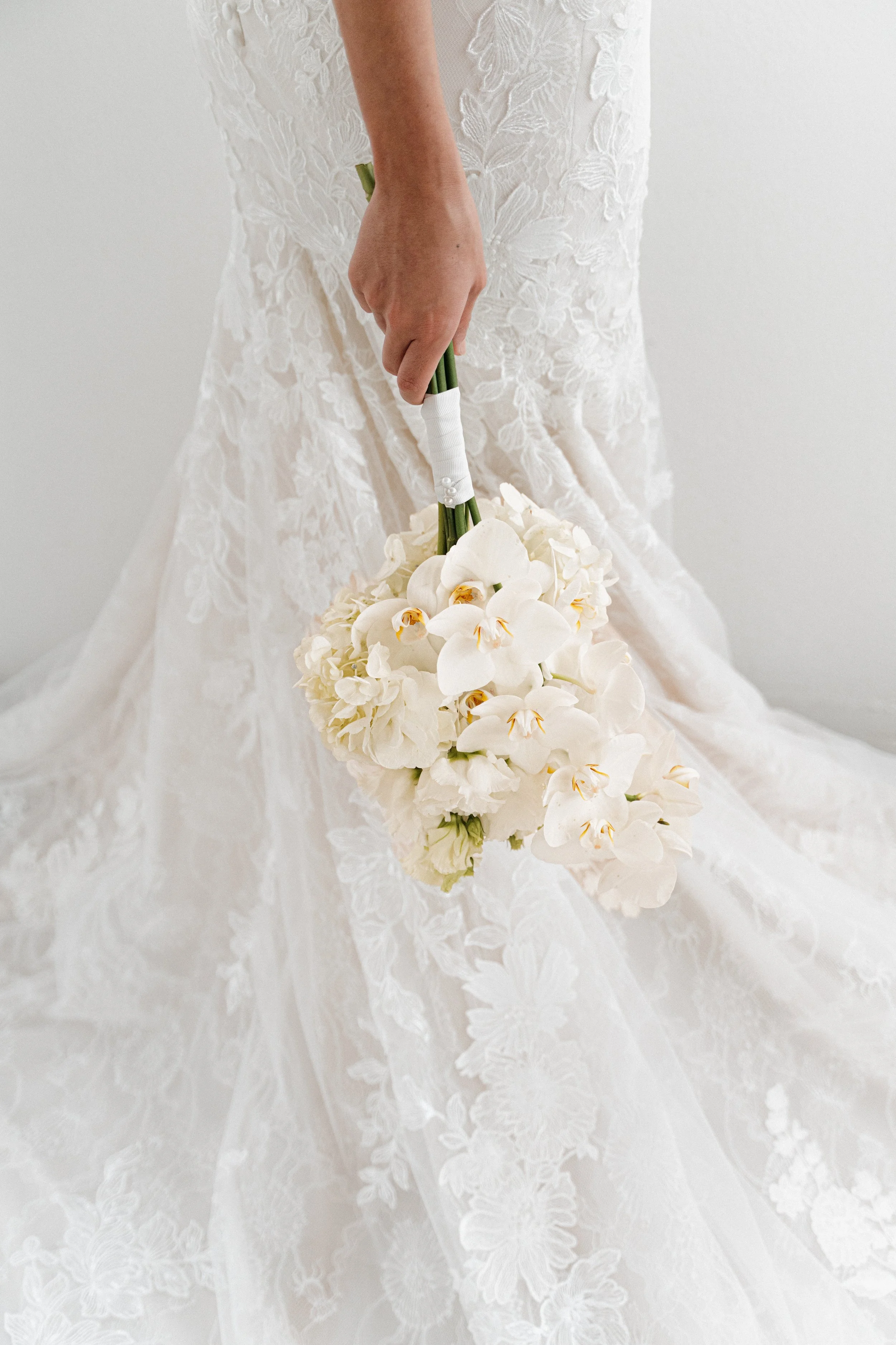Une mariée en robe de mariage en dentelle blanche, tenant un bouquet de fleurs blanches dont des orchidées, dans une pièce au fond blanc.