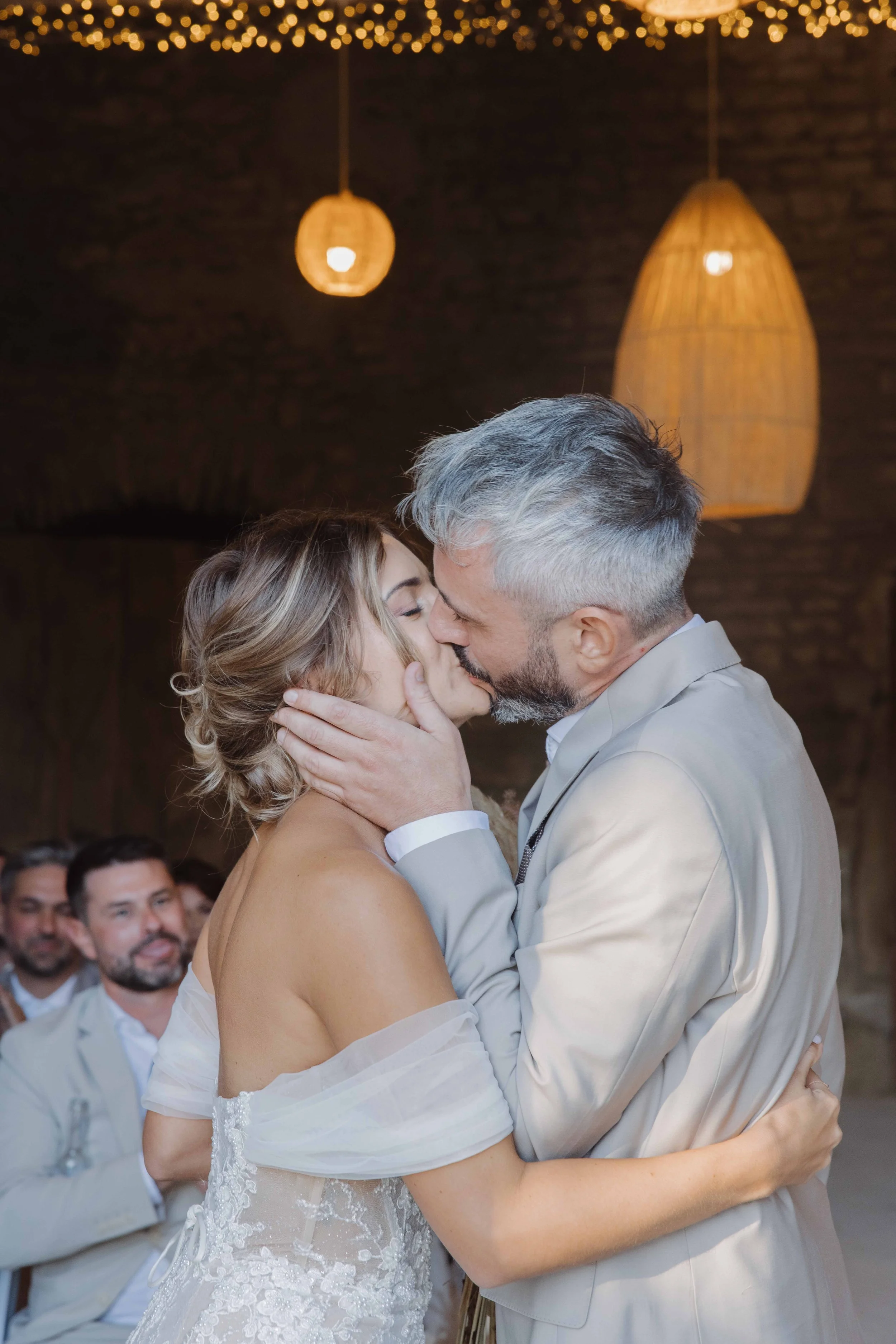 Un couple échange un baiser lors d'un mariage, entouré de proches assis. L'intérieur est décoré de lampes suspendues en rotin et de guirlandes lumineuses. Photographe mariage Isère, ferme du rocher, cérémonie civil et laïque. Bordeaux et Lyon.