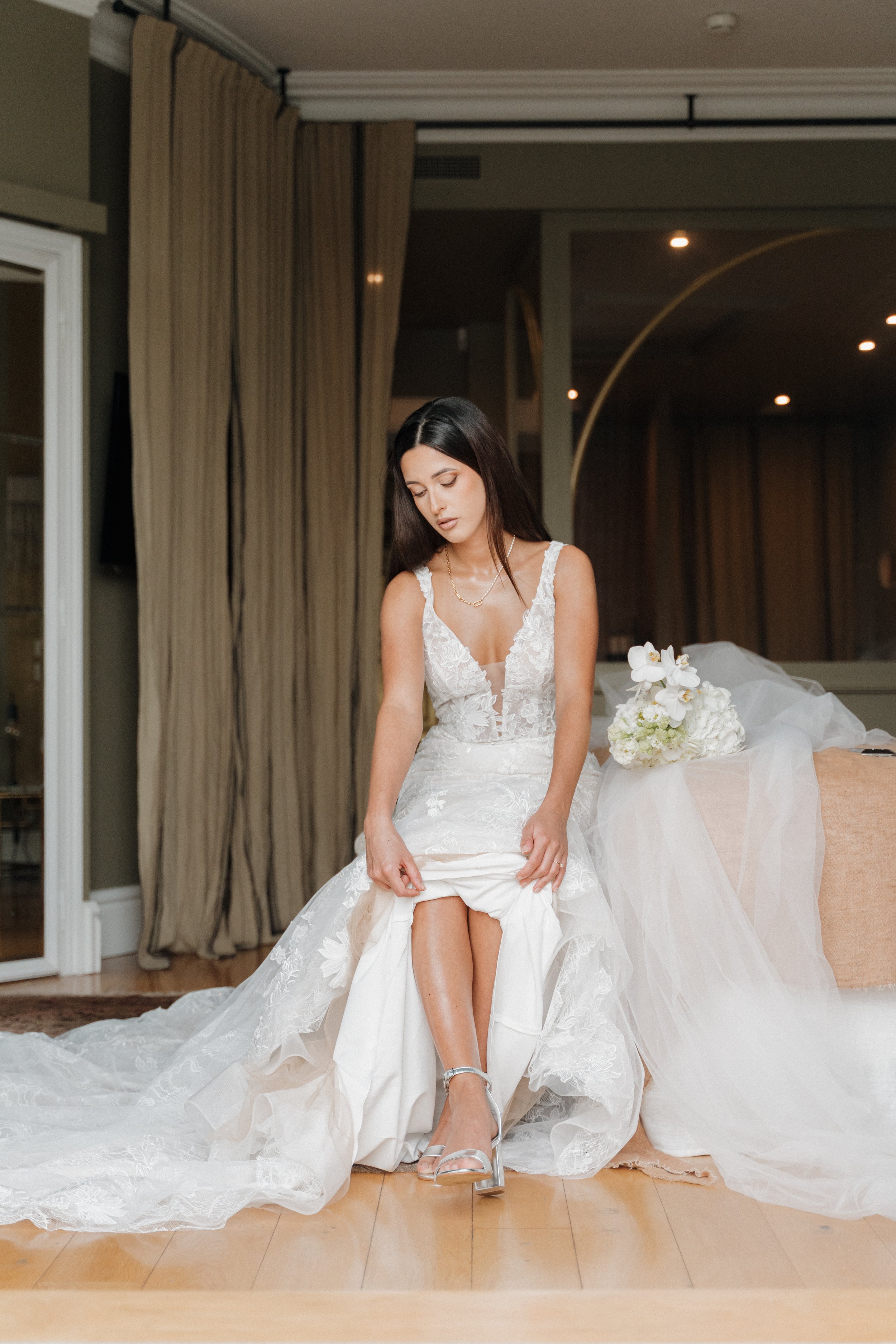 Une jeune femme en robe de mariage, assise en levant légèrement sa robe, dans une pièce élégante avec des rideaux beige et un bouquet de fleurs blanches à côté.  session femme mariées et robes haut de gamme, brindos, hôtel et lac,  photographe 