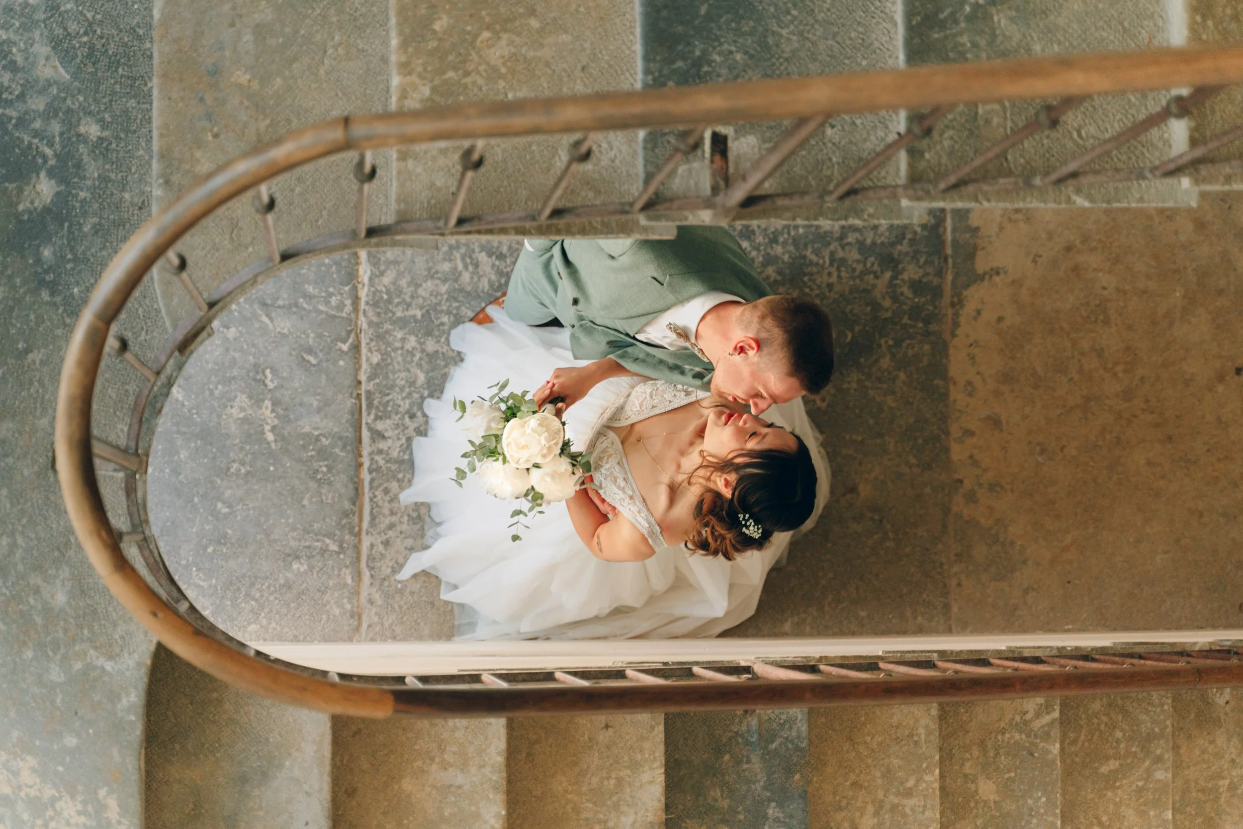 Un couple de mariés s'embrasse sur un escalier en pierre, la femme porte une robe de mariée blanche et tient un bouquet de roses blanches, l'homme porte un costume vert pale. Domaine de tourieux à Savigny dans le beaujolais à Lyon. Mariage cérémonie 