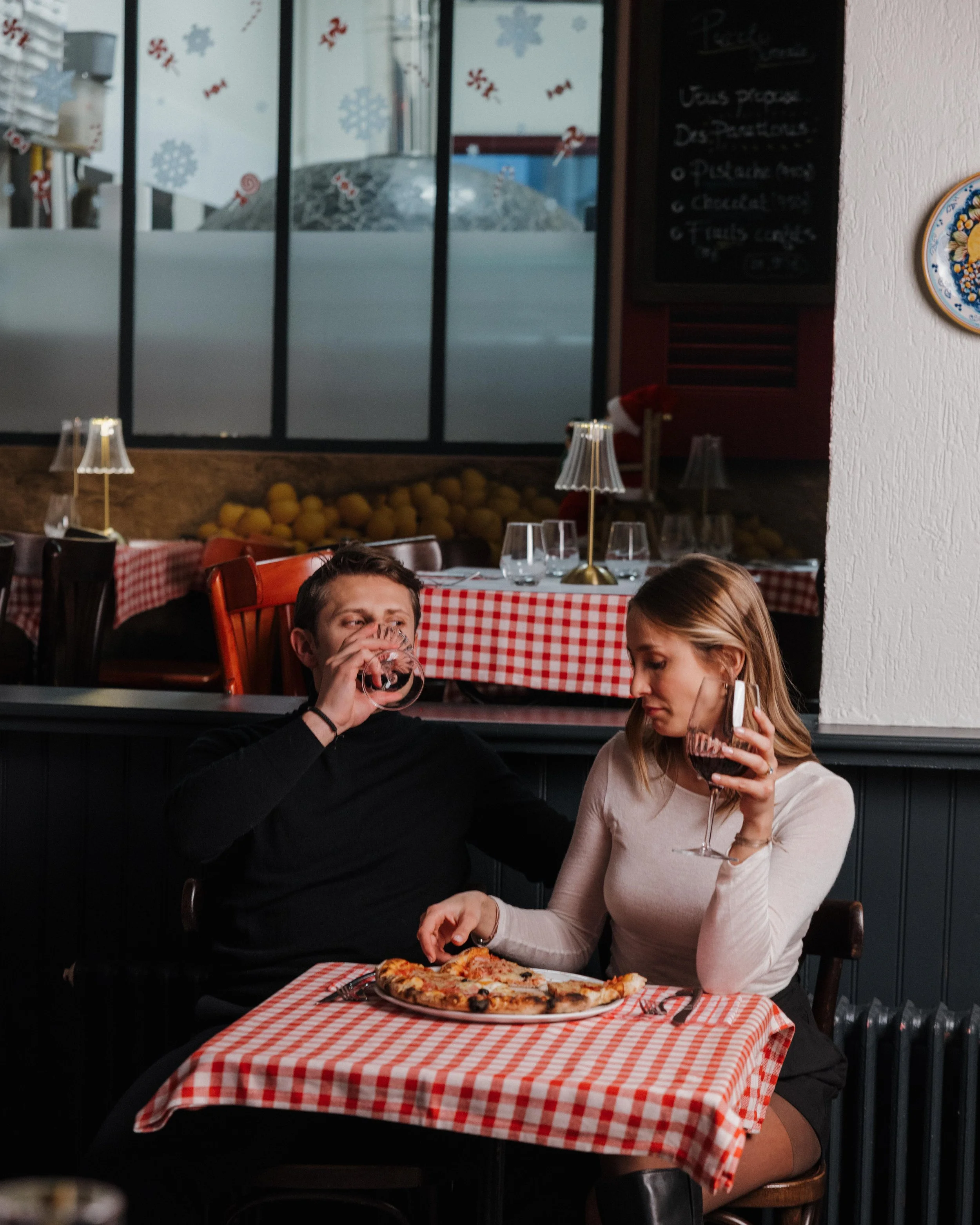 Un homme et une femme assis à une table dans un restaurant, partageant une pizza et buvant du vin rouge session engagement couple Lyon 6eme, brotteaux, photographe mariage bordeaux et Lyon et destination wedding 