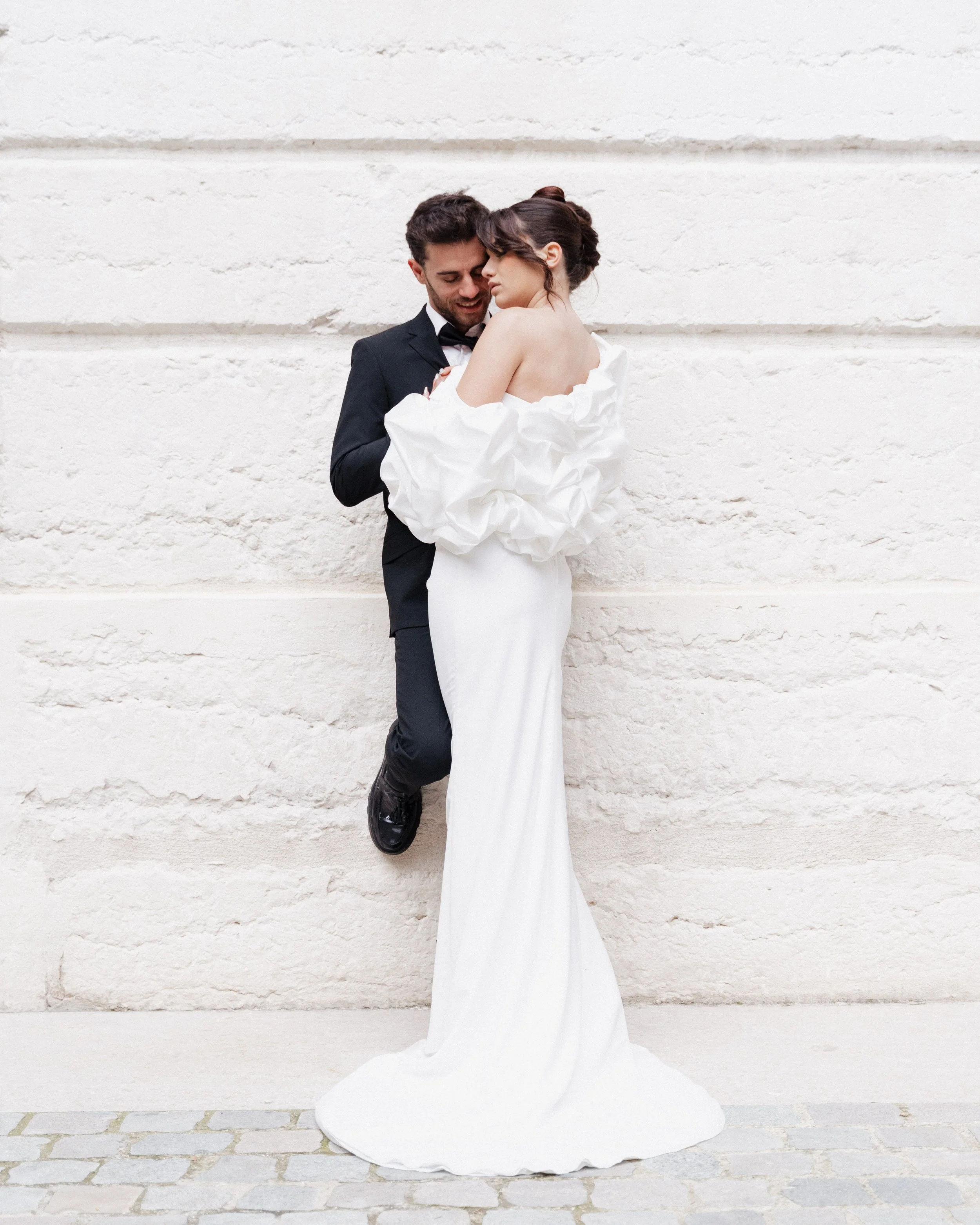 Un couple en costume de mariage posant contre un mur en pierre, la femme en robe blanche élégante et le homme en costume noir, avec une ambiance romantique à Lyon 2ème à l'hôtel dieu à l'intercontinental lors d'une session engagement.