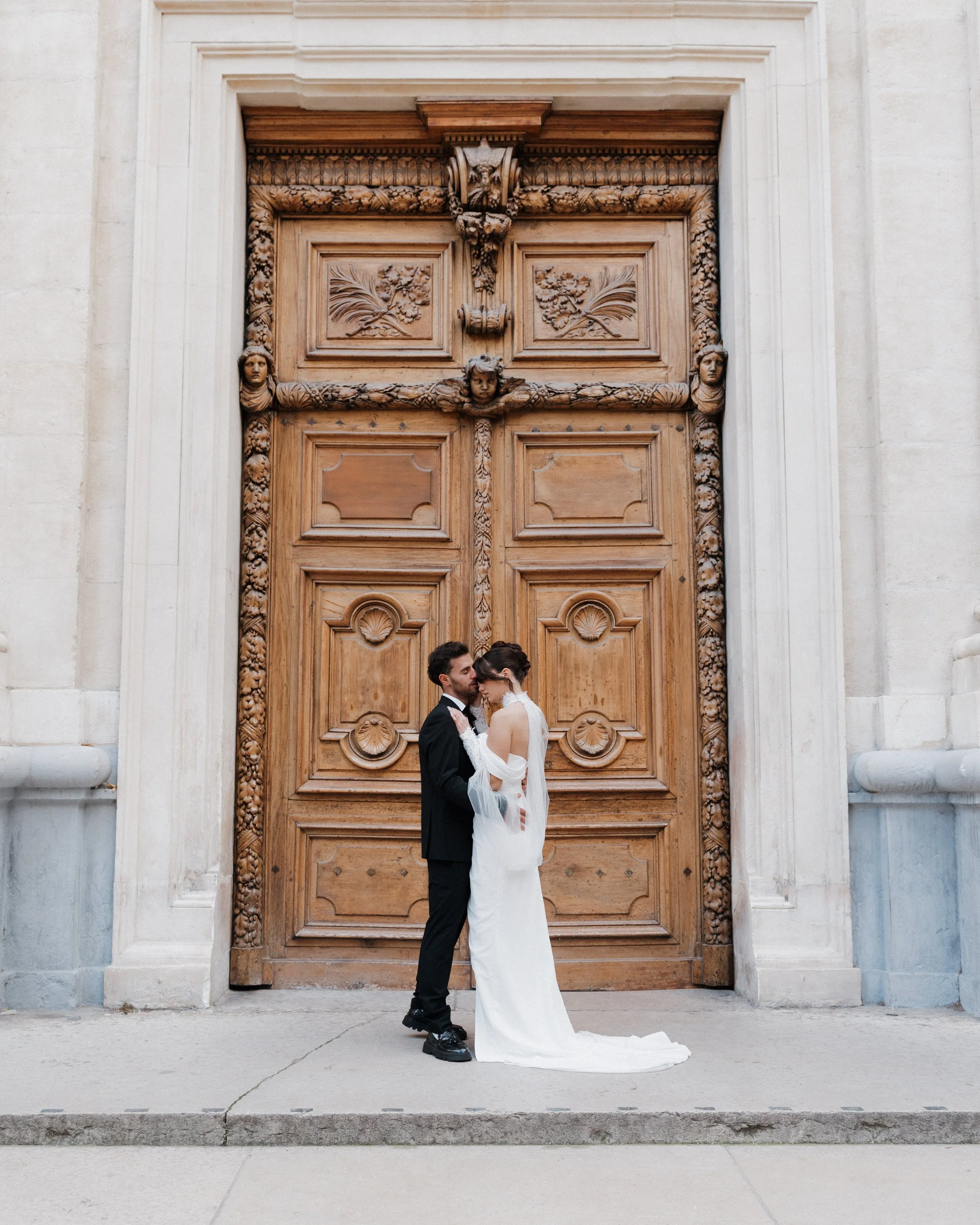 Un couple en vêtements de mariage devant une grande porte en bois avec des sculptures sculptées, probablement devant une église ou un bâtiment historique à Lyon 2ème à l'hôtel dieu à l'intercontinental 