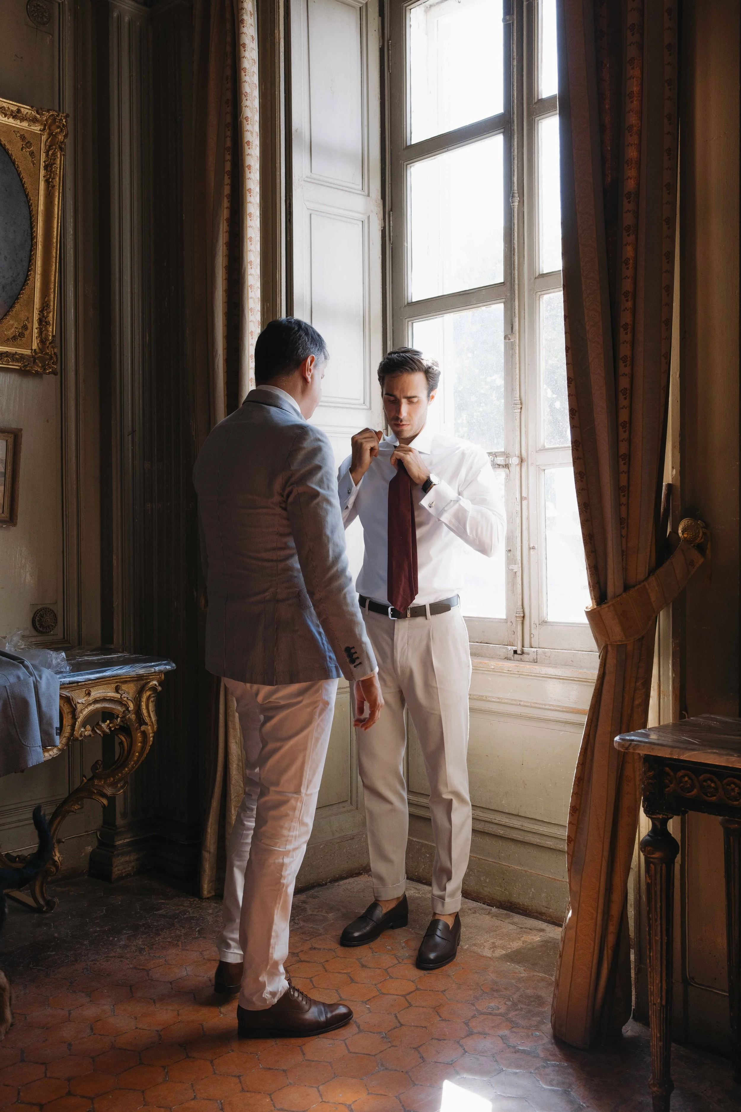 Deux hommes habillés en costume blanc et beige, ajustant une cravate dans une pièce ornée de meubles anciens, près d'une grande fenêtre avec des rideaux marron. Mariage au chateau de lachal dans le rhone alpes à côté de Lyon. Cérémonie civil 