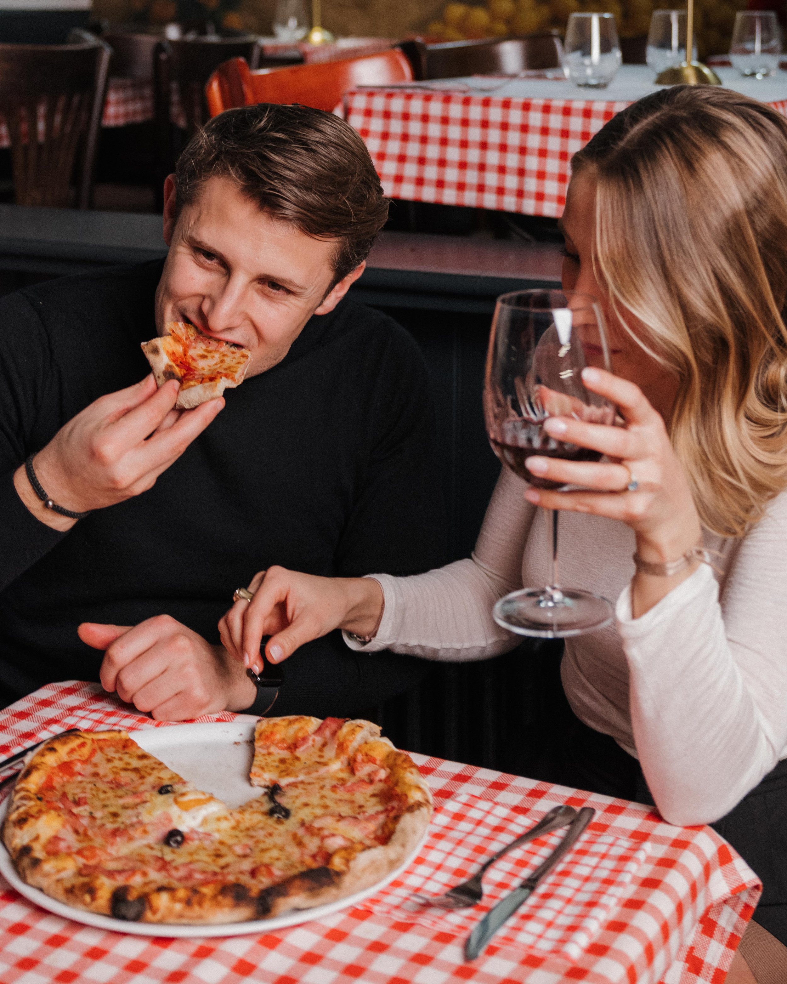 Un couple mangeant une pizza dans un restaurant, la femme sirote du vin rouge, la table est couverte d'une nappe à carreaux rouges et blancs. session engagement couple Lyon 6eme, brotteaux, photographe mariage bordeaux et Lyon et destination wedding