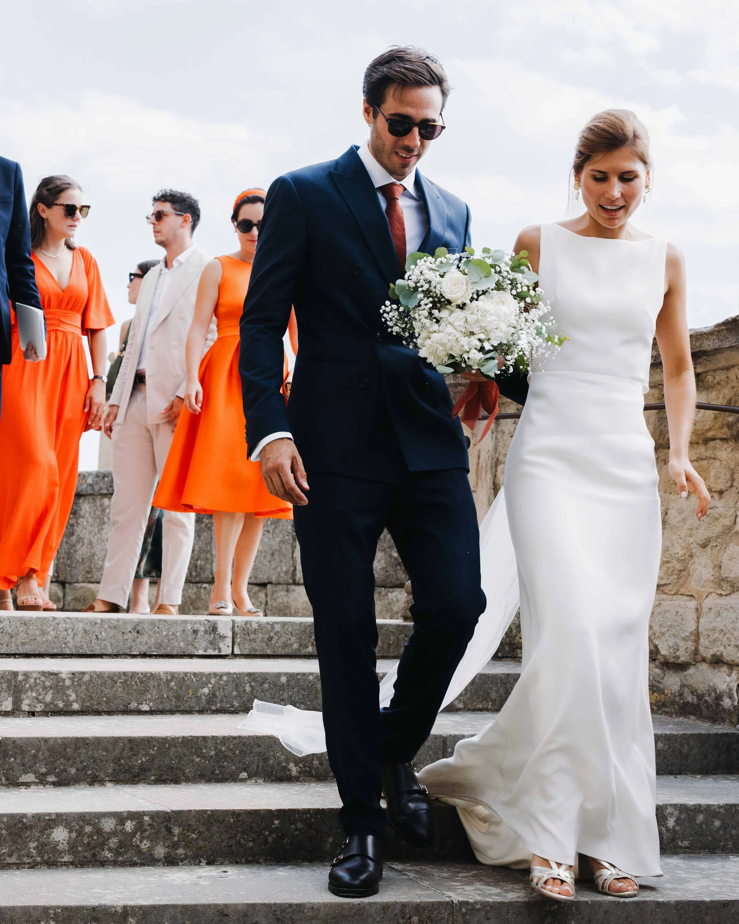 Un couple de mariés descend des escaliers lors de leur mariage, entouré de leurs invités, avec le ciel nuageux en arrière-plan. Domaine du bijoutier à grignan, drome provençal, coucher de soleil, session couple, mariage religieux, cérémonie 