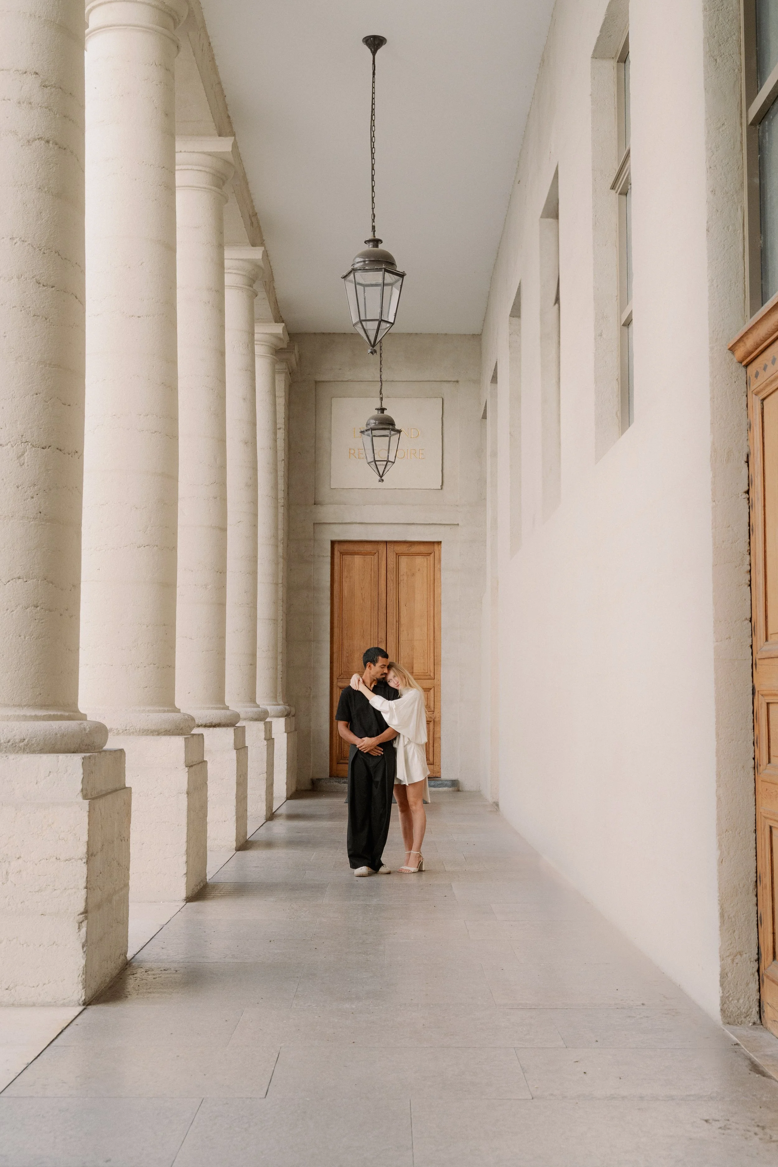 Un couple se tient dans une galerie avec de grandes colonnes en pierre et un plafond haut, partageant un moment intime à l'hôtel dieu à Lyon 2ème, à l'intercontinental, session engament, photographe mariage duo haut de gamme.