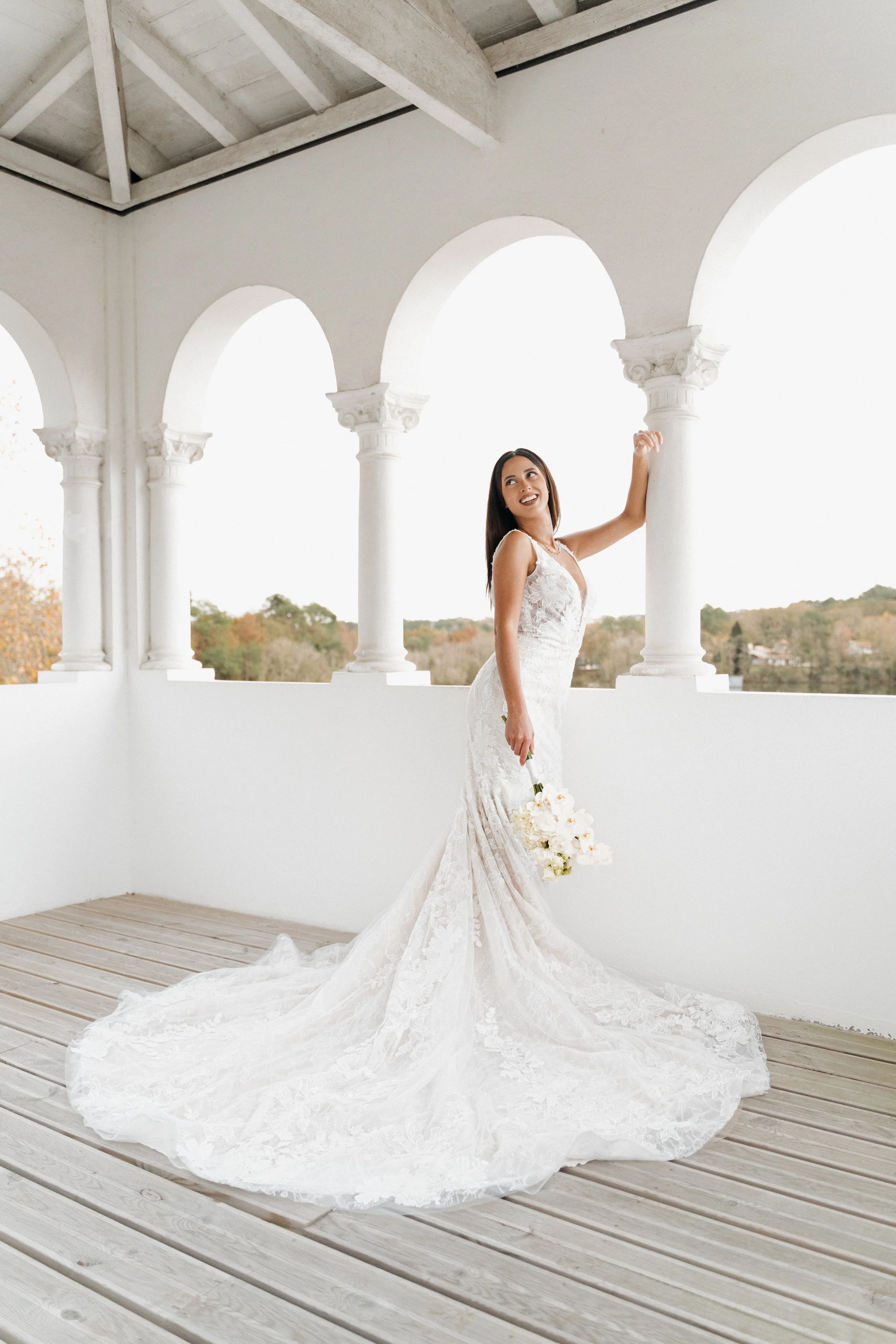 Jeune femme en robe de mariée blanche dans une structure avec colonnes blanches, tenant un bouquet de fleurs blanches, souriant et regardant vers la droite, décor naturel en arrière-plan.  session femme mariées et robes haut de gamme, brindos, hôtel 