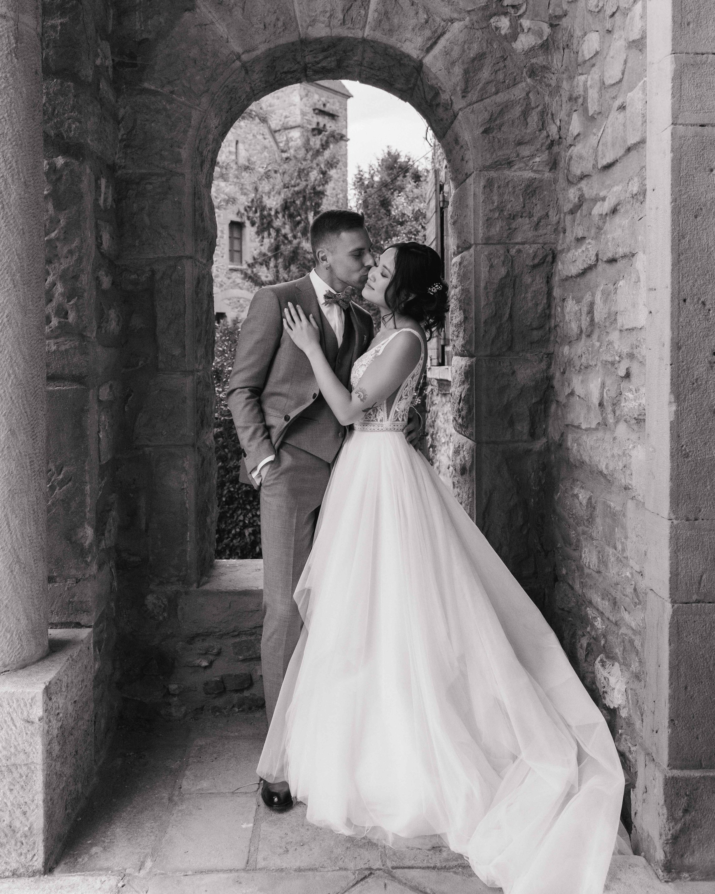 Un couple de mariés s'embrassant sous une arche en pierre, dans un style noir et blanc. Domaine de tourieux à Savigny dans le beaujolais à Lyon. Mariage cérémonie laïque, photographe Lyon haut de gamme, duo photographe, argentique.