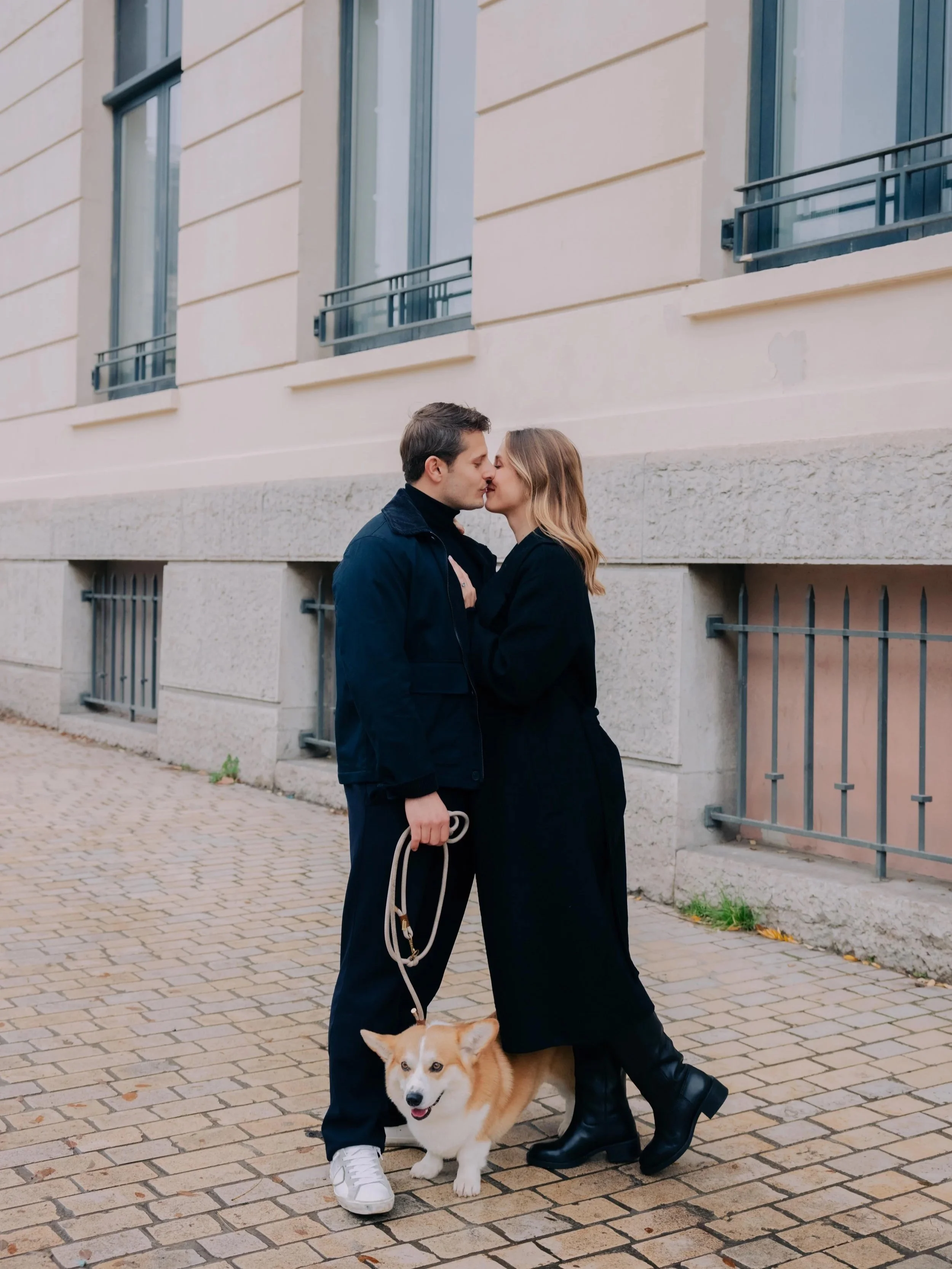 Un couple s'embrassant sur un trottoir pavé, tenant la laisse d'un chien corgi, devant un bâtiment résidentiel.