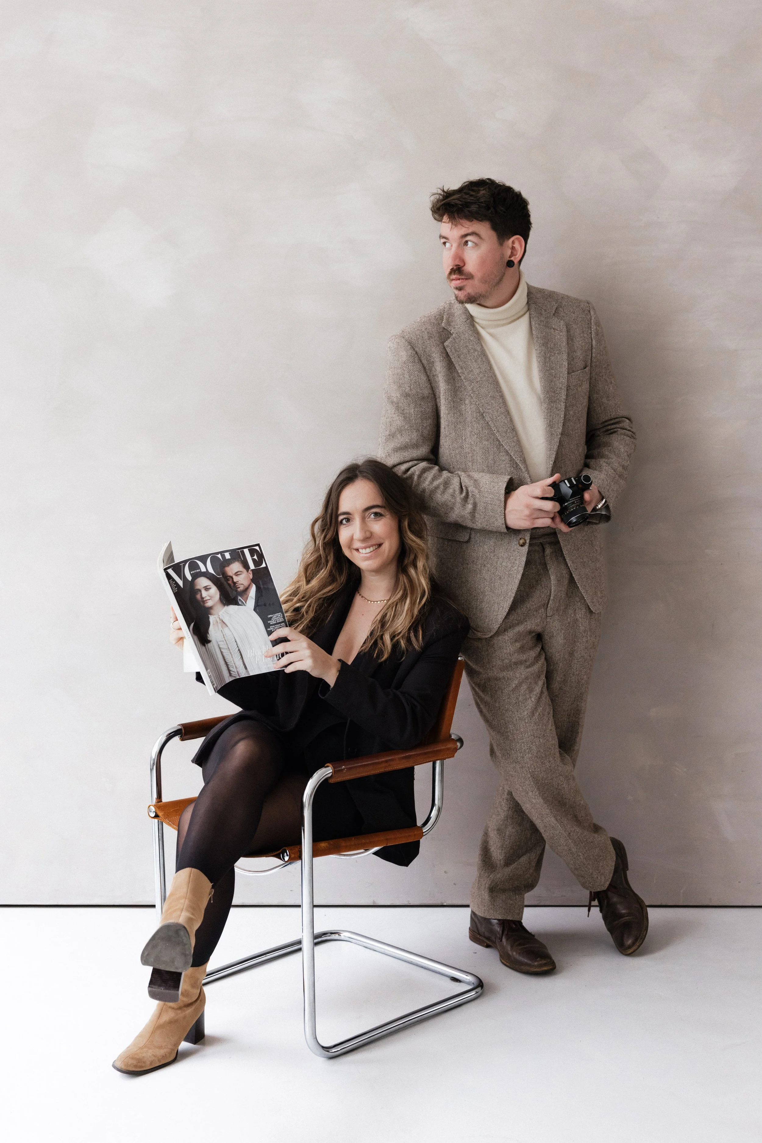 Une femme assise sur une chaise en bois et métal tenant un magazine Vogue, et un homme debout à côté d'elle tenant un appareil photo, tous deux dans un environnement minimaliste avec un mur beige clair en arrière-plan. Photographe Lyon, bordeaux