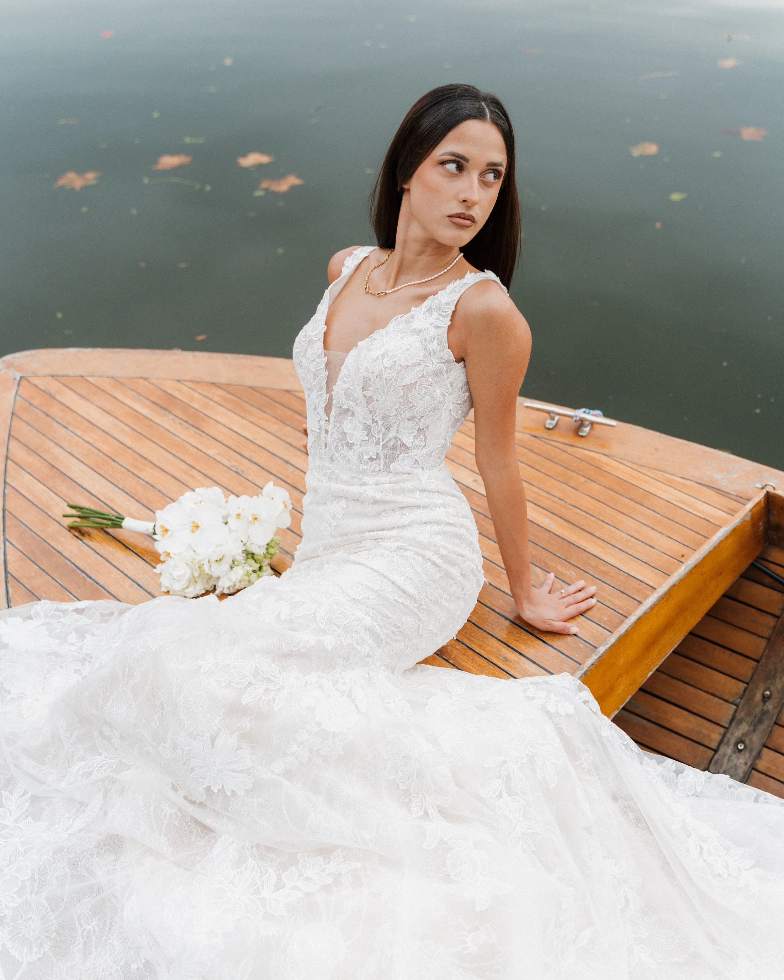 Une femme en robe de mariée blanche, assise sur une embarcation en bois au bord de l'eau, avec un bouquet de fleurs blanches à côté d'elle.  session femme mariées et robes haut de gamme, brindos, hôtel et lac,  photographe mariage bordeaux et Lyon 