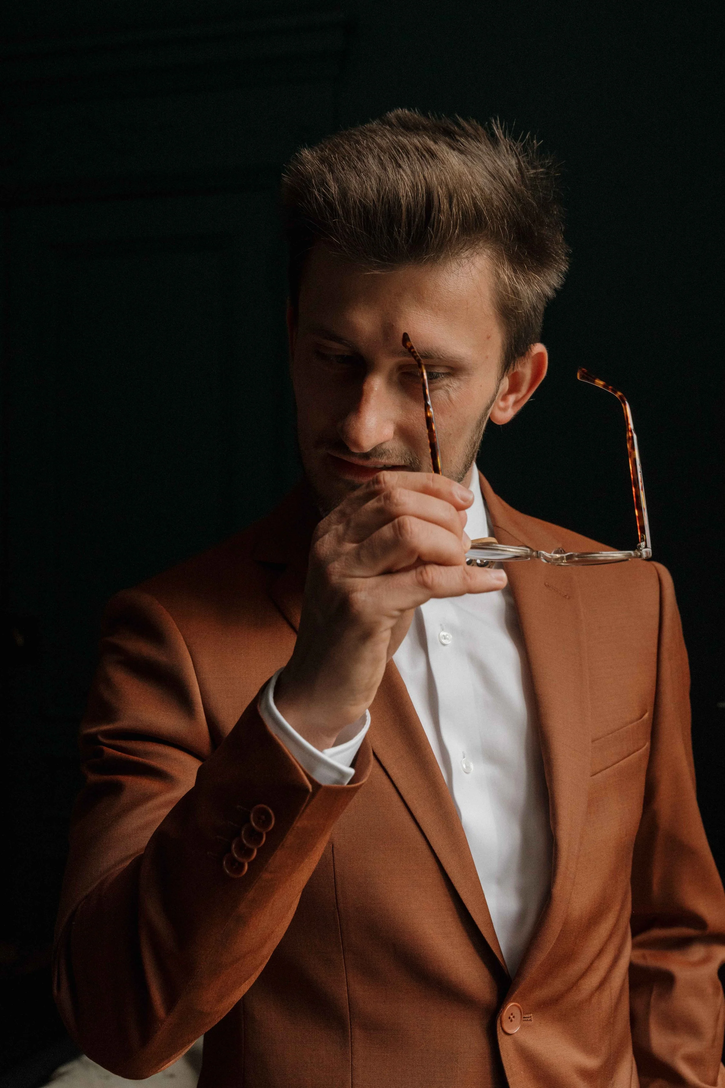 Un homme en costume marron tient ses lunettes en regardant vers le bas, contre un fond sombre. Domaine de tourieux à Savigny dans le beaujolais à Lyon. Mariage cérémonie laïque, photographe Lyon haut de gamme, duo photographe, argentique.