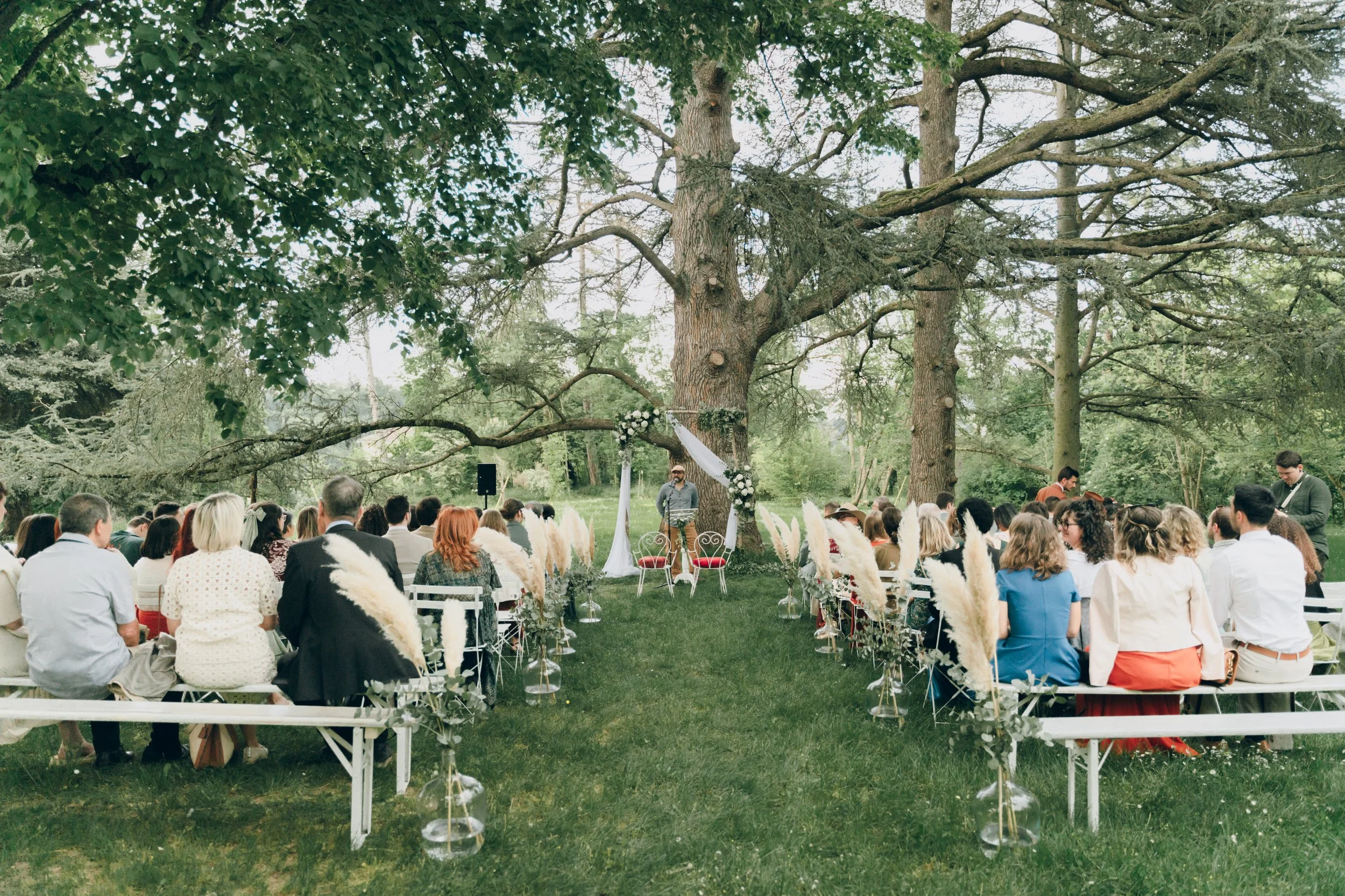 Cérémonie de mariage en plein air sous un grand arbre avec des invités assis, décoration florale et arce décoratif au centre.  Domaine de tourieux à Savigny dans le beaujolais à Lyon. Mariage cérémonie laïque, photographe Lyon haut de gamme, film 