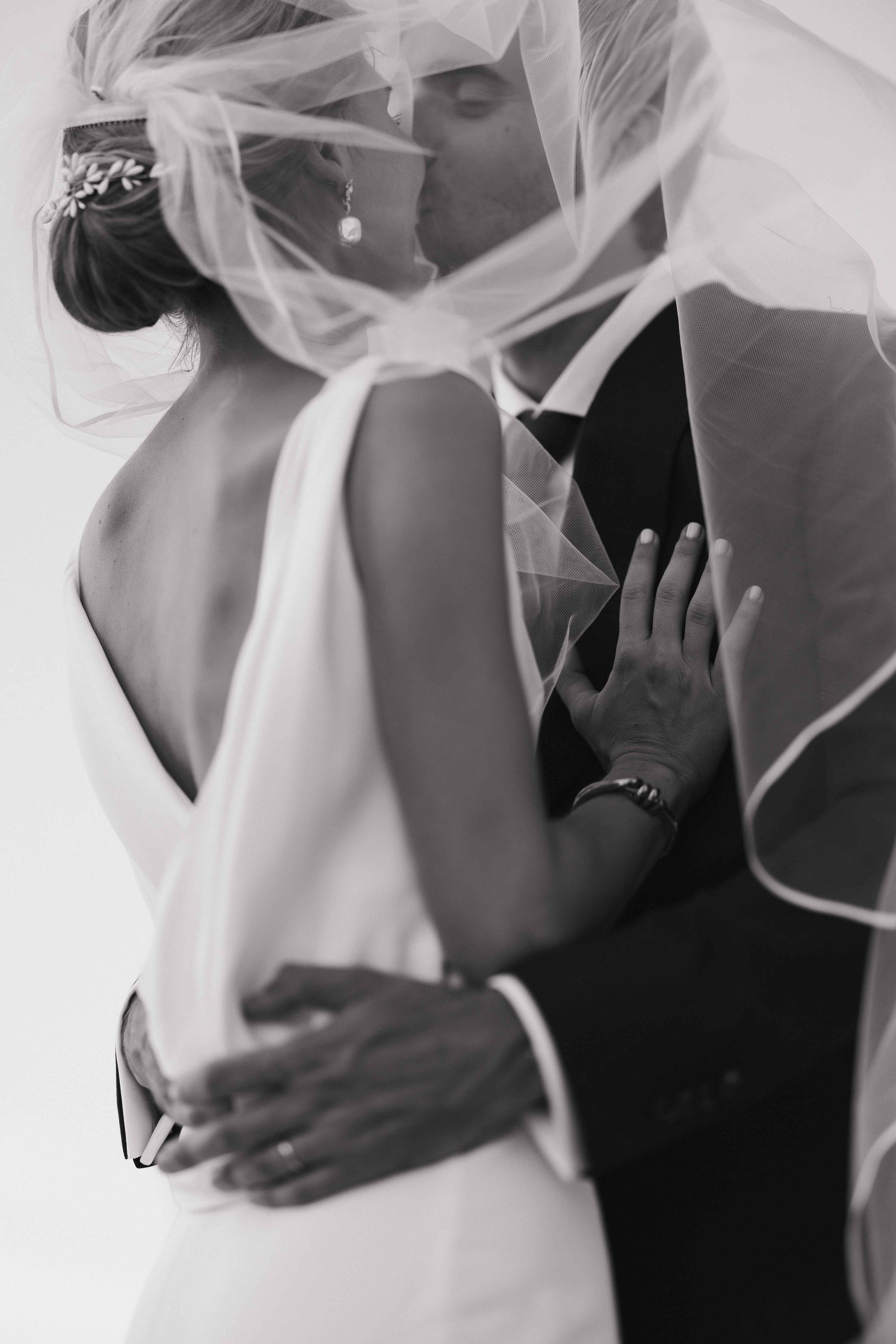 Un couple de mariés s'embrassant et se tenant par la taille, enveloppés partiellement par un voile blanc dans une photo en noir et blanc. Photo session couple, grignan, drome provençal, session engagement, domaine du bijoutier
