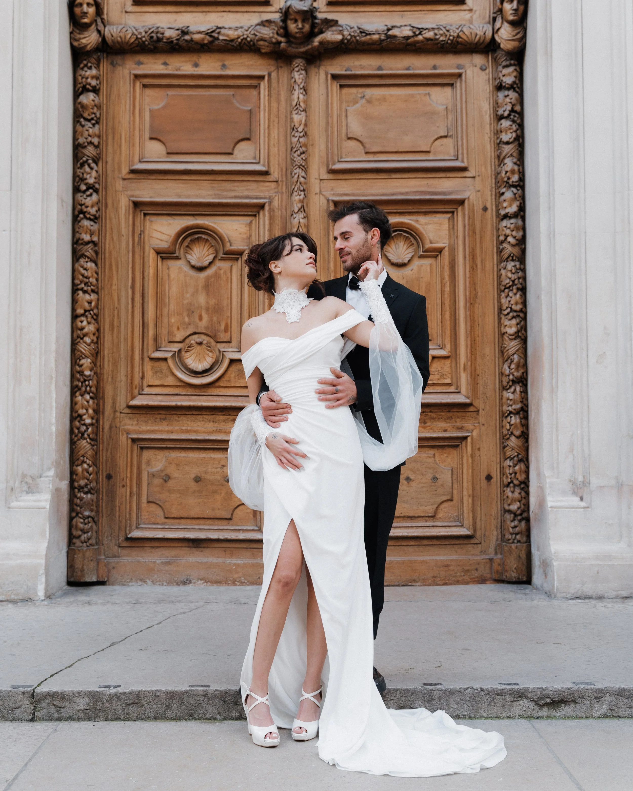 Un couple en mariage posant devant une grande porte en bois, lui en costume noir et elle en robe blanche élégante à l'hôtel dieu à Lyon 2ème à l'intercontinental 