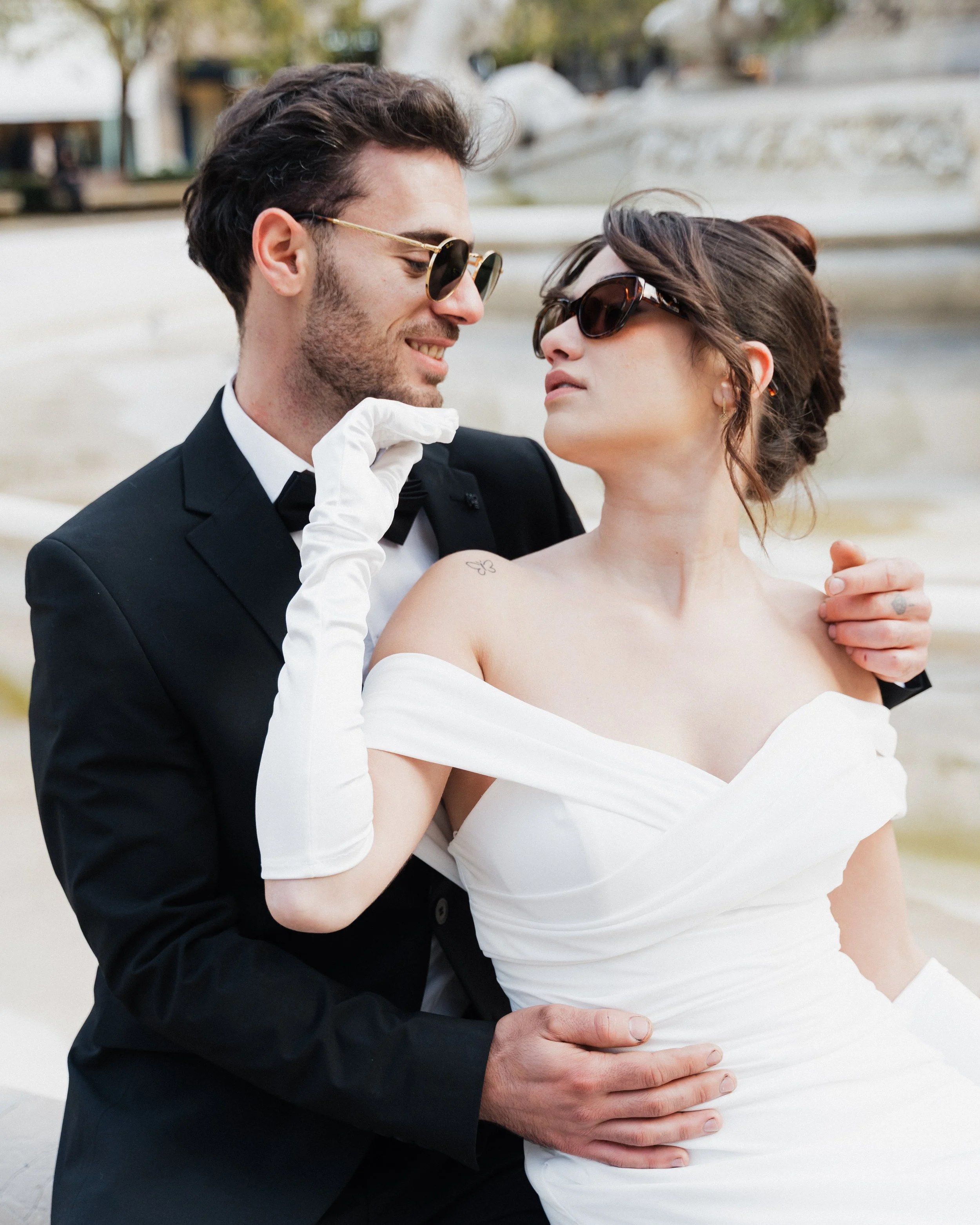 Un couple habillé en vêtements élégants, en compagnie d'une fontaine ou d'un monument, se regarde de près. La femme porte une robe blanche et des lunettes de soleil, tandis que l'homme porte un costume noir et des lunettes de soleil. La scène évoque 