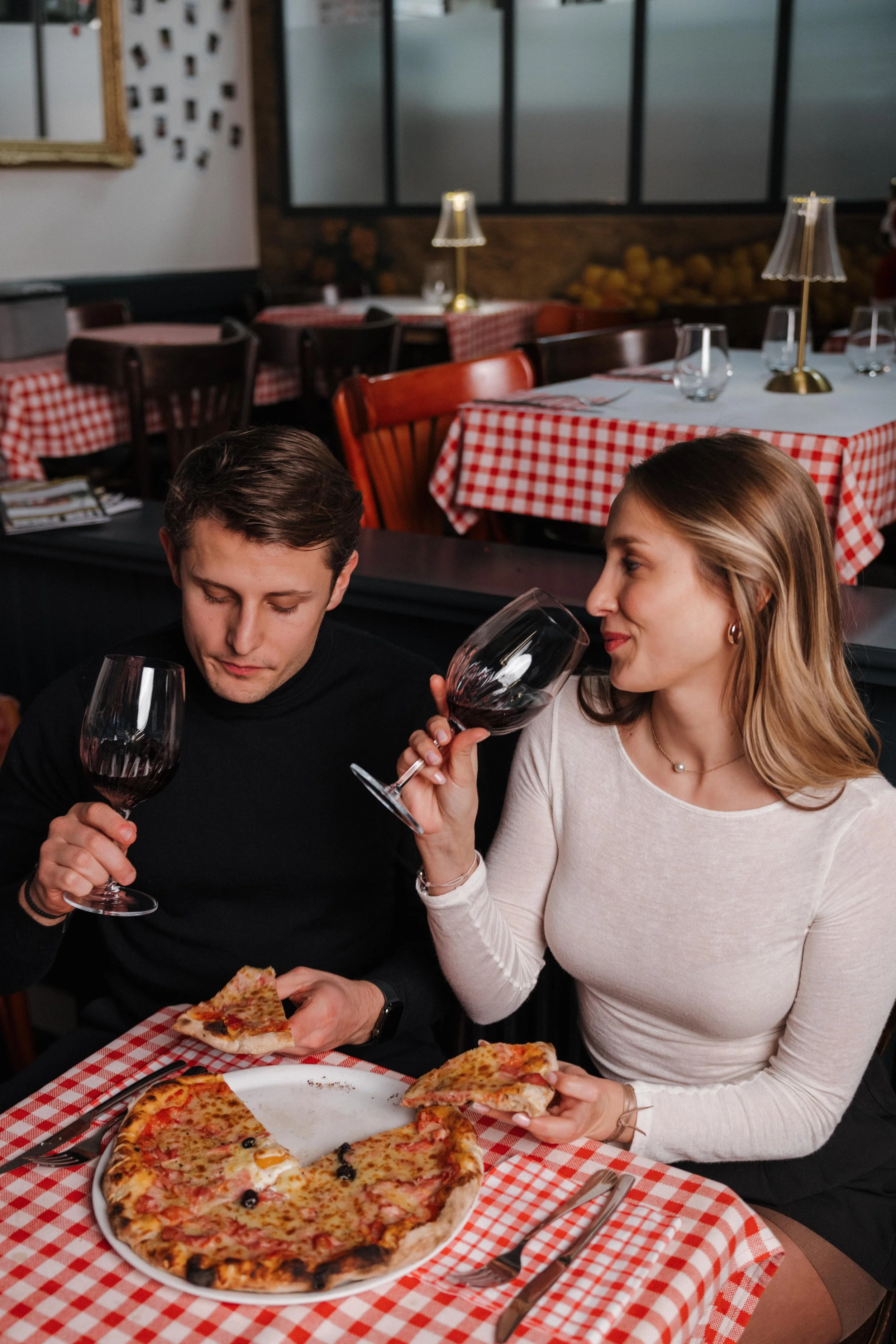 Deux personnes dégustent une pizza et du vin dans un restaurant avec une décoration à carreaux rouges et blancs, session engagement couple Lyon 6eme, brotteaux, photographe mariage bordeaux et Lyon et destination wedding