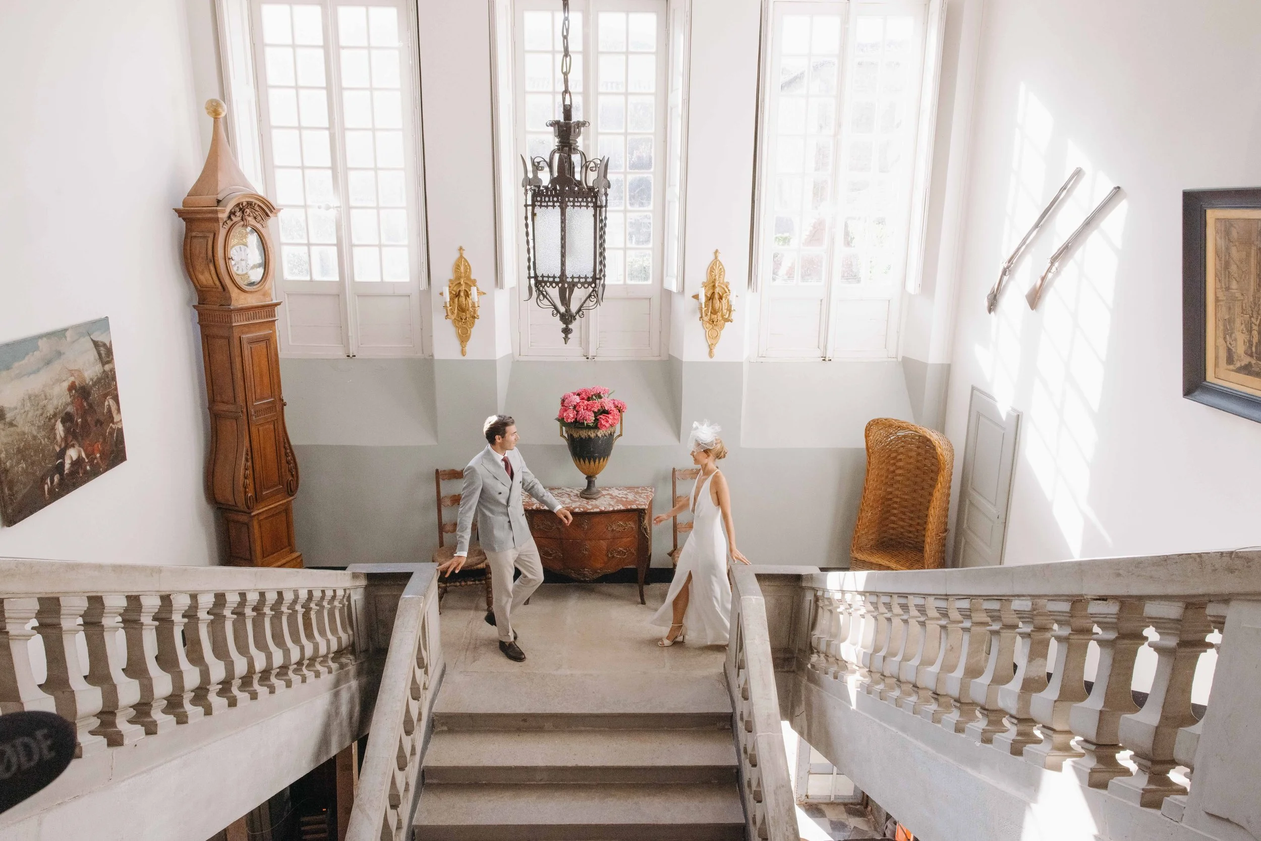 Un couple habillé pour un mariage se tenant par la main dans un intérieur élégant avec un escalier en marbre, un grand vase de fleurs roses, une horloge ancienne, et une cheminée en bois. Mariage au chateau de lachal dans le rhone alpes.