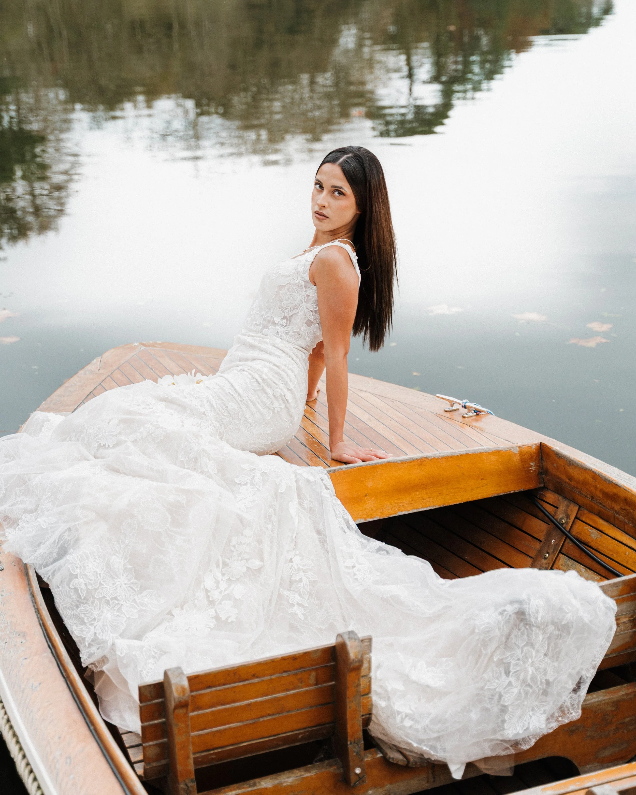 Jeune femme en robe de mariée assise à l'arrière d'un bateau en bois sur un lac calme, réfléchissant dans l'eau.  session femme mariées et robes haut de gamme, brindos, hôtel et lac,  photographe mariage bordeaux et Lyon et destination wedding