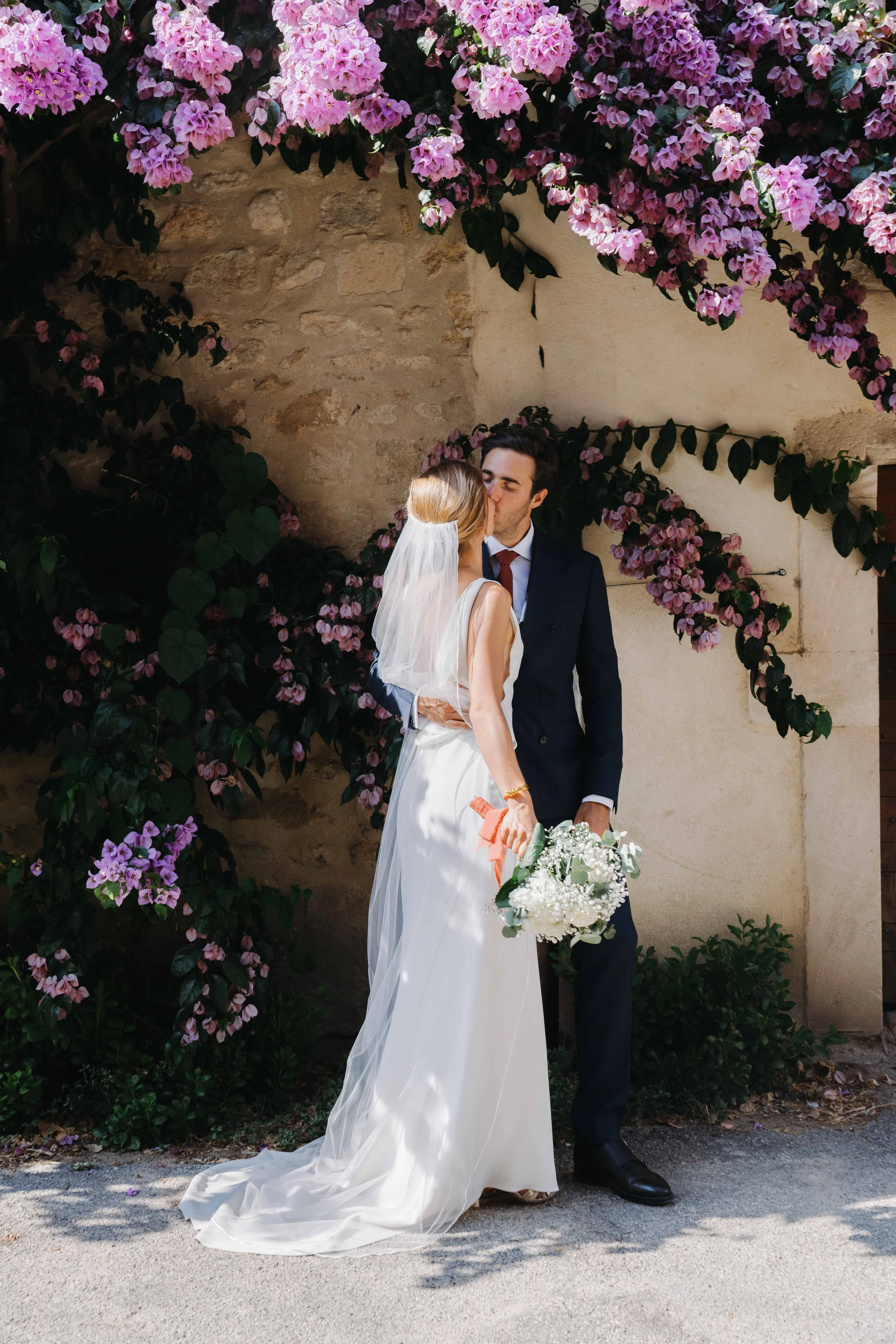 Un couple de mariés s'embrassant devant un mur de pierres recouvert de fleurs de bougainvilliers roses, lors d'un mariage en plein air. Domaine du bijoutier à grignan, drome provençal, coucher de soleil, session couple, mariage religieux, 
