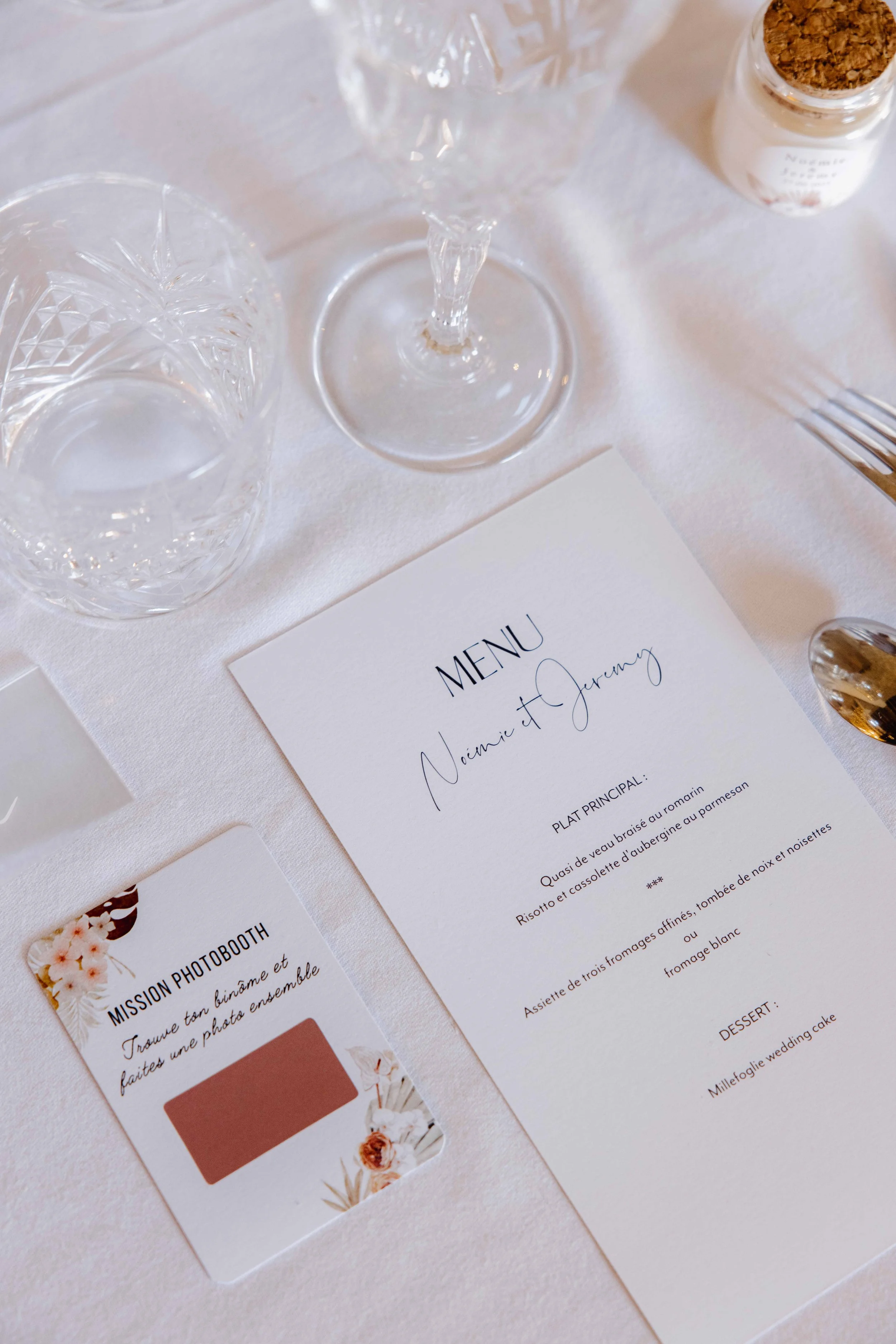 Carte de menu de mariage avec eau, verre à vin et à eau en cristal, petit pot à épices et carte de mission photographique sur une table blanche. Photographe mariage Isère, ferme du rocher, cérémonie civil et laïque. Bordeaux et Lyon. 