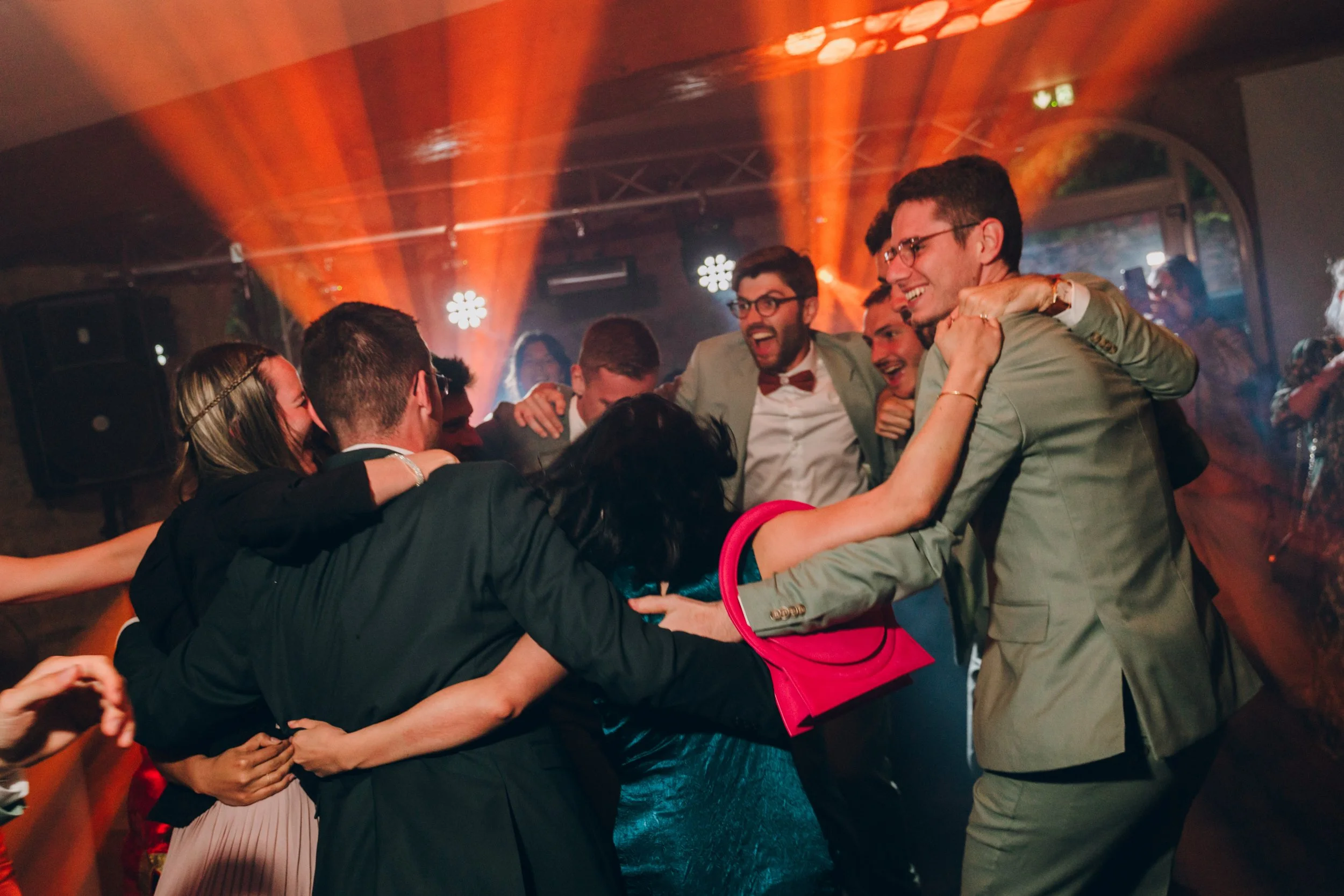 Groupe de personnes vêtu de façon formelle, qui danse et s'amuse lors d'une fête ou d'un événement, avec des lumières colorées en arrière-plan. Domaine de tourieux à Savigny dans le beaujolais à Lyon. Mariage cérémonie laïque, photographe Lyon 