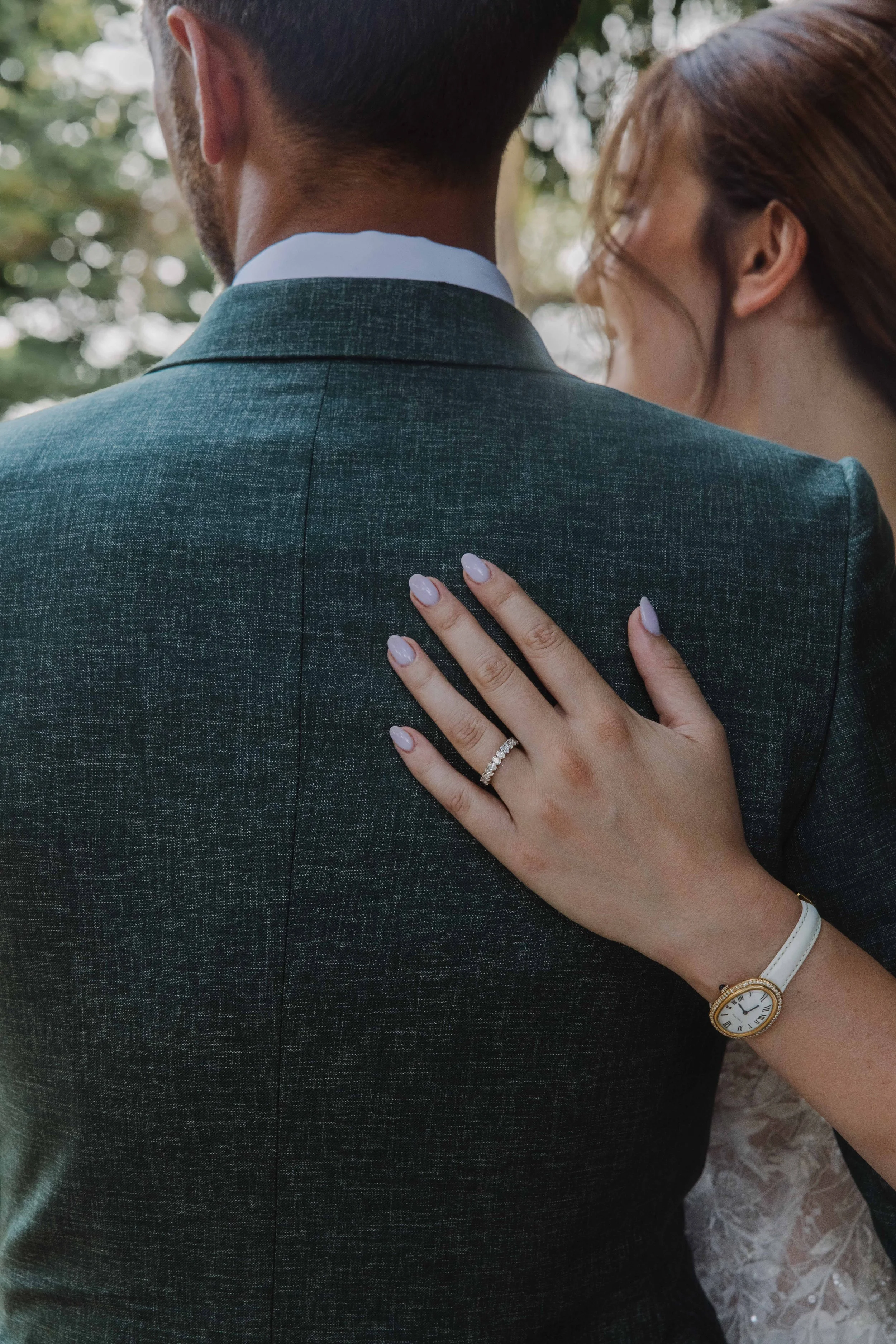 Close-up d'une personne portant une montre blanche et une bague sur l'annulaire, posée sur l'épaule d'un homme en costume gris foncé, lors d'une cérémonie ou d'un moment intime en extérieur. Mariage chateau de champ renard, Blacé, beaujolais, Lyon