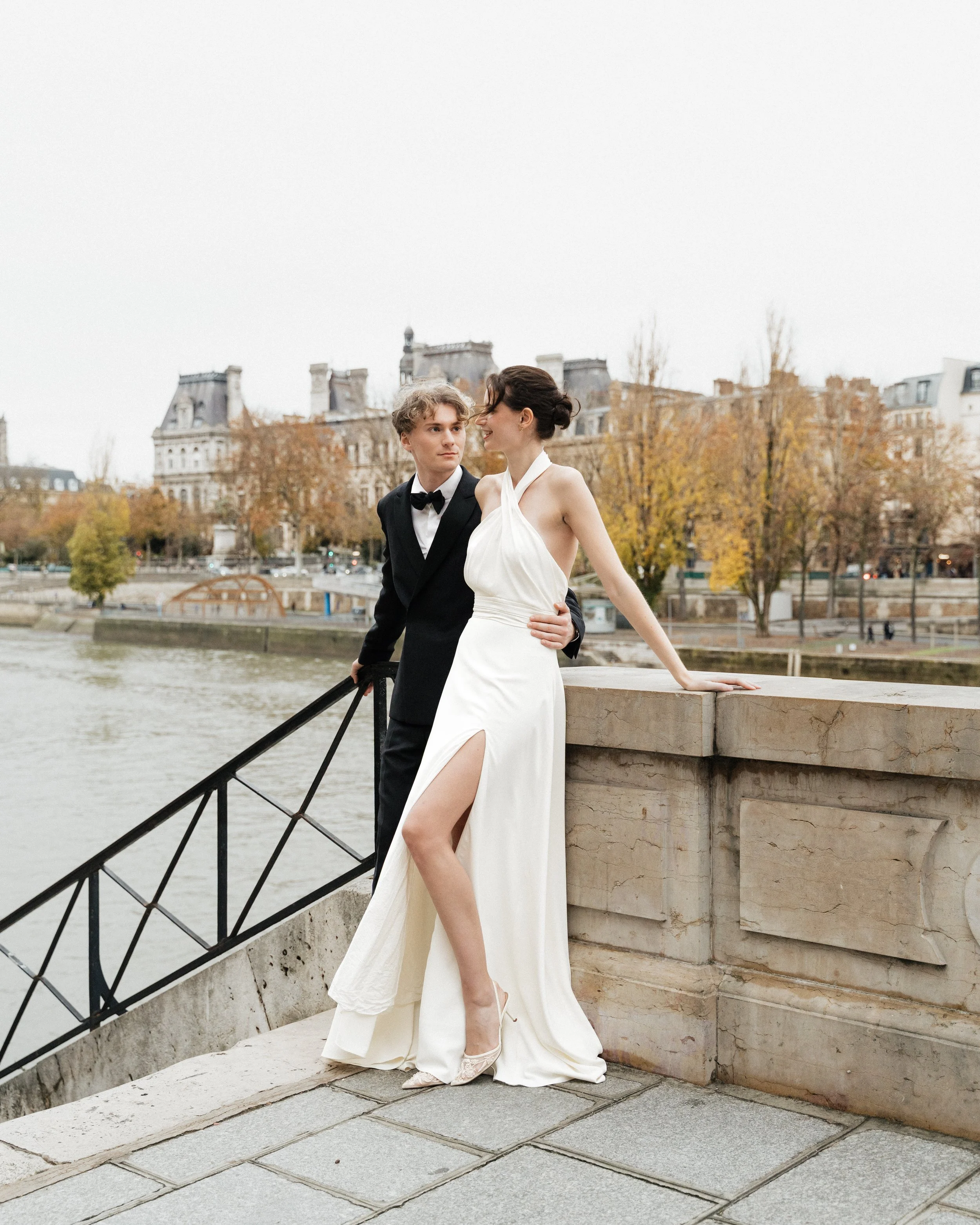 Un couple en robe de mariée et costume se tient près d'une rivière à Paris lors d'une journée automnale. Session engagement paris, Louvre, mariage haut de gamme, destination wedding. Robe de mariées, madame a dit oui