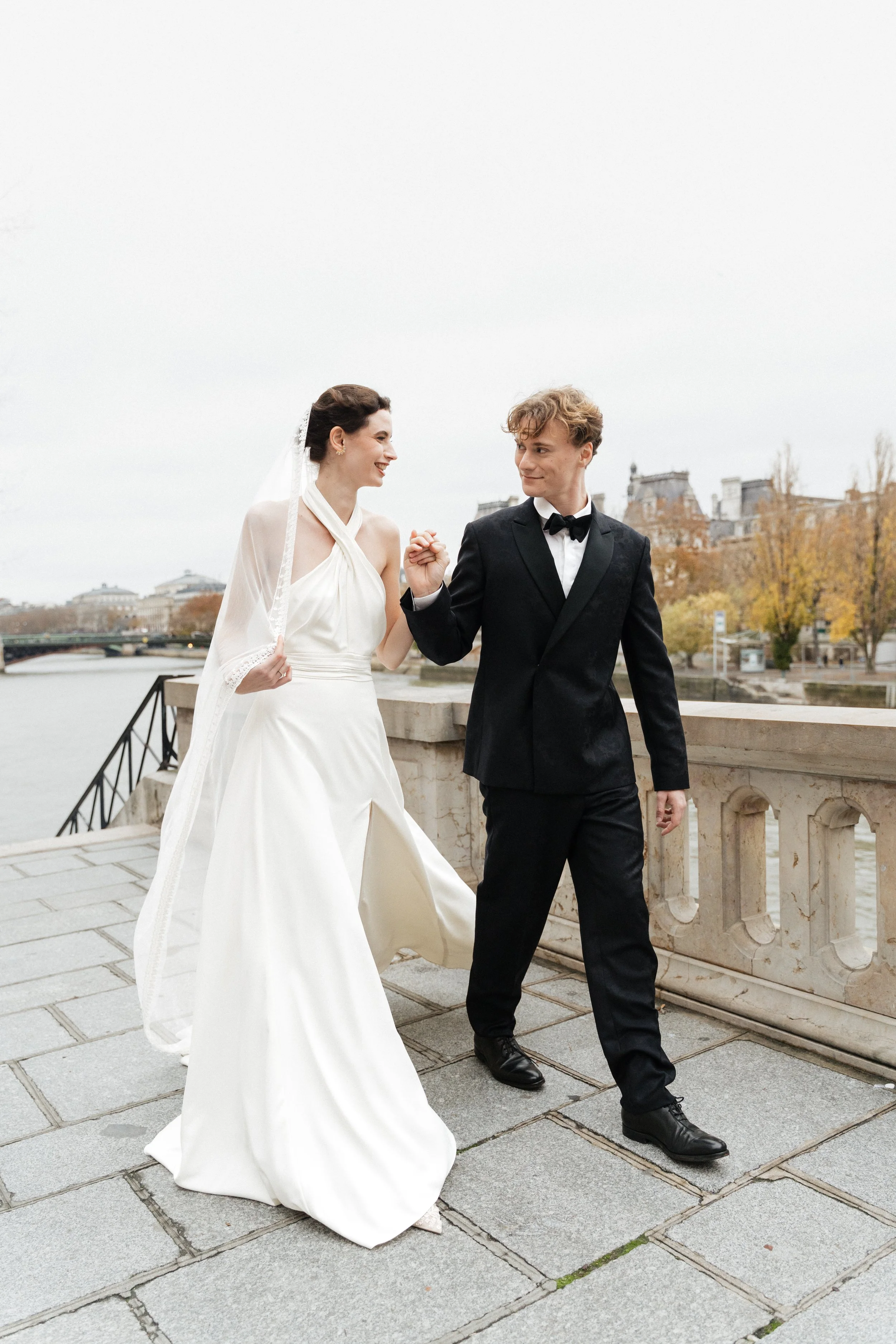 Un couple de mariés tenant la main, marchant sur un pont au bord de la Seine à Paris, avec des bâtiments historiques en arrière-plan. Session engagement paris, Louvre, mariage haut de gamme, destination wedding. Robe de mariées, madame a dit oui