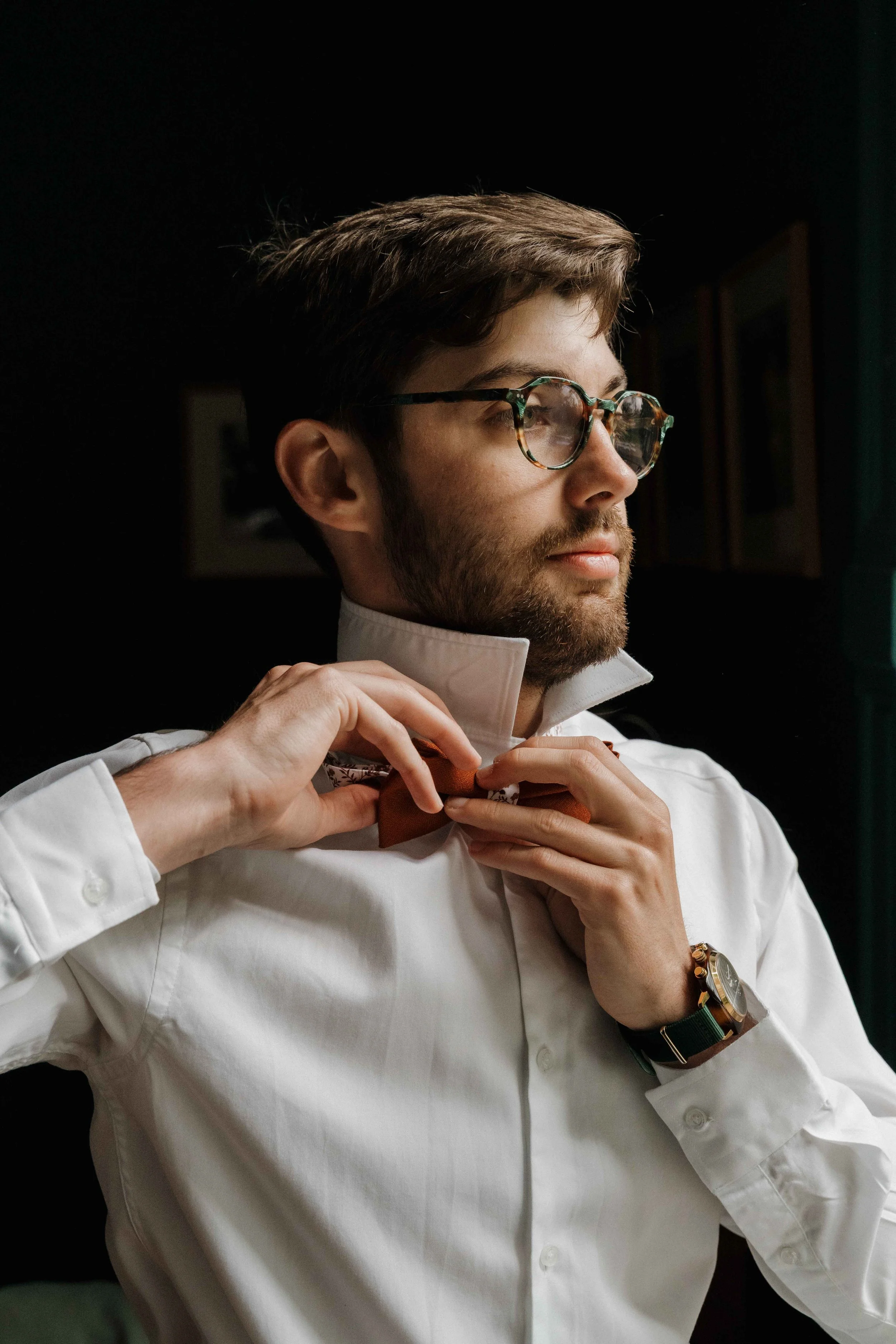 Un homme avec une barbe, portant des lunettes à monture tachetée, ajuste un nœud papillon orange en satin, habillé d'une chemise blanche. Domaine de tourieux à Savigny dans le beaujolais à Lyon. Mariage cérémonie laïque, photographe Lyon haut de gamm