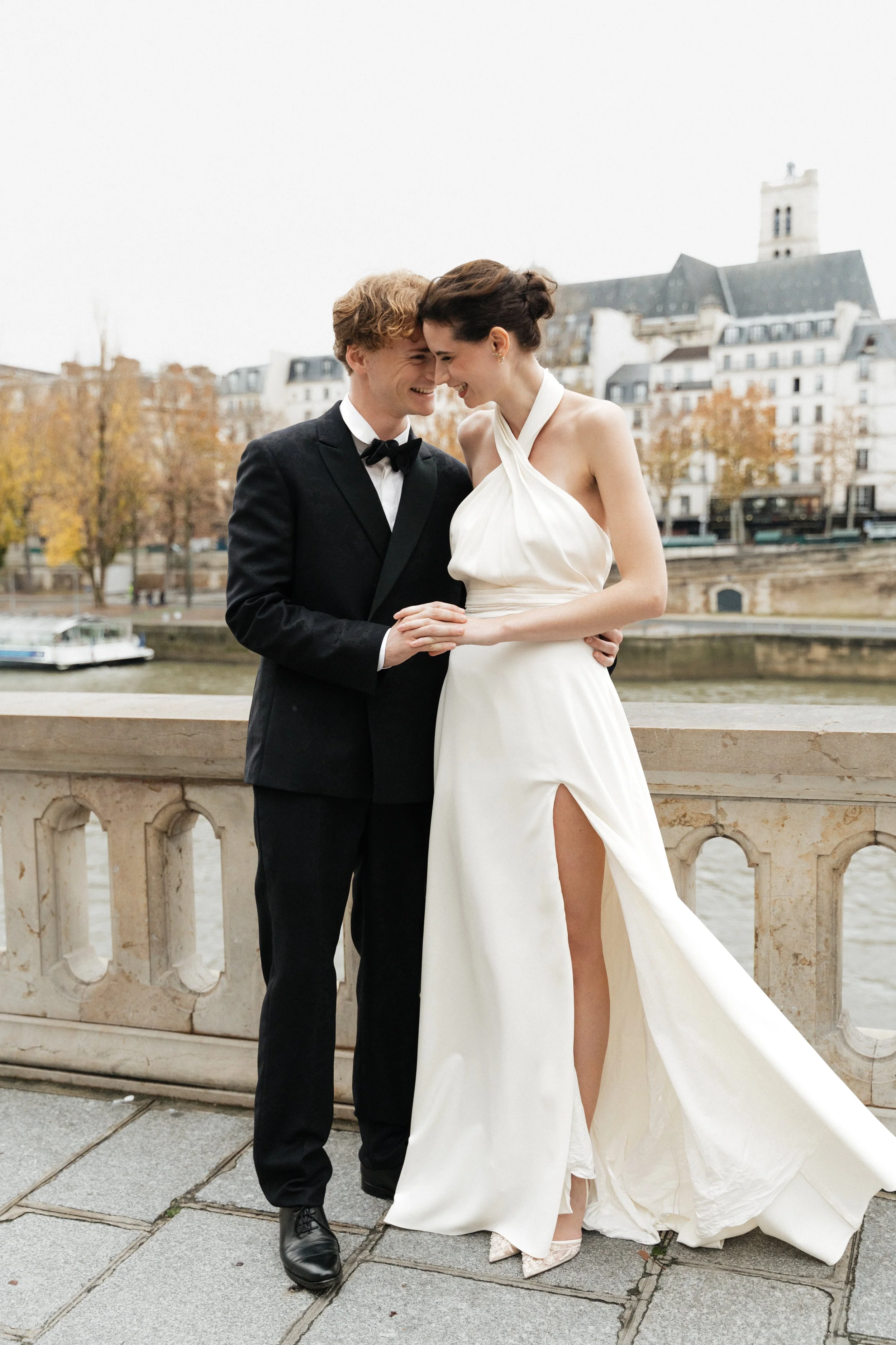 Un couple de mariés souriants, en vêtements de mariage, échangeant un regard dans un décor urbain au bord de la Seine, à Paris. Session engagement paris, Louvre, mariage haut de gamme, destination wedding. Robe de mariées, madame a dit oui