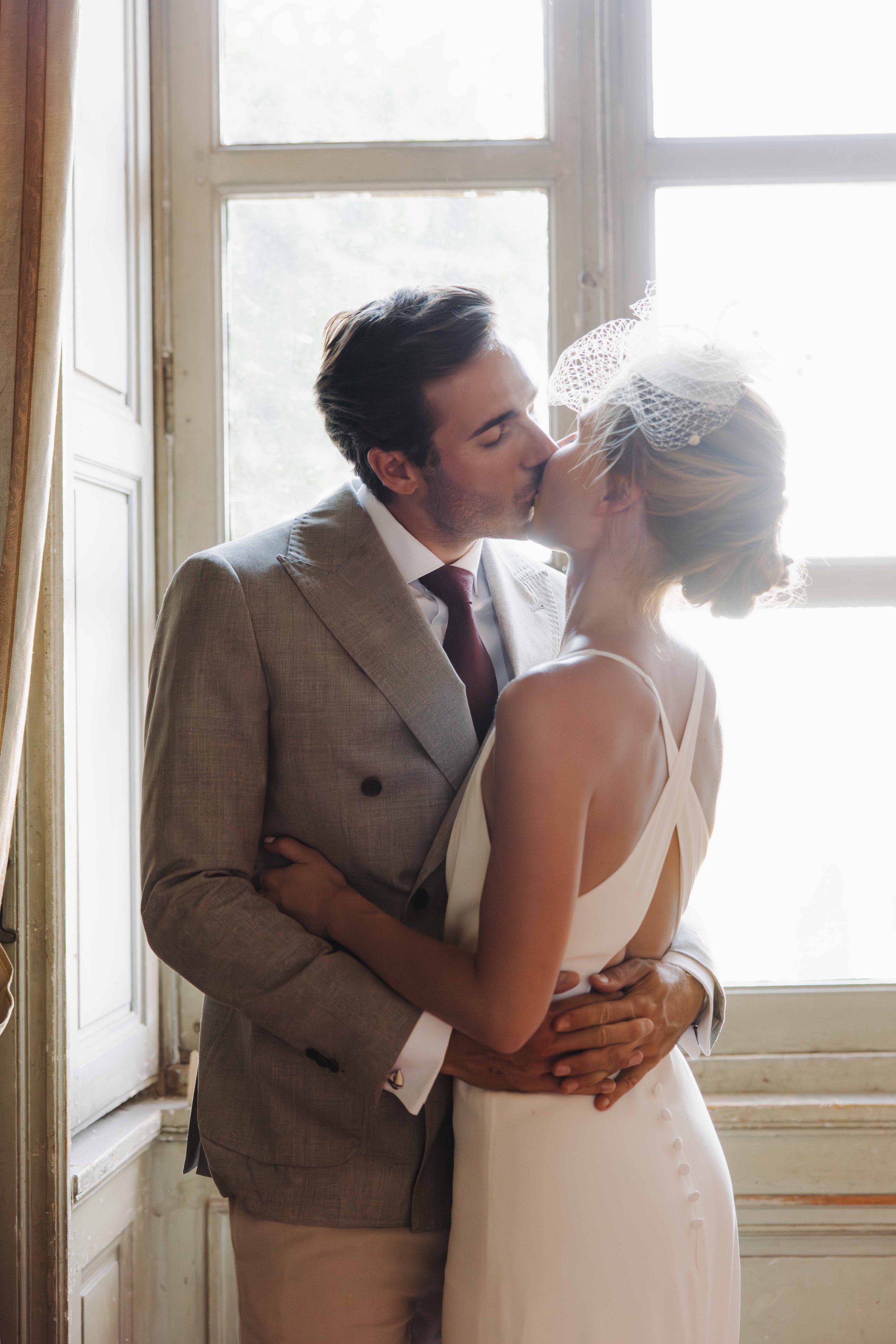 Un couple en habits de mariage s'embrassant devant une fenêtre lumineuse. Mariage au chateau de lachal dans le rhone alpes à côté de Lyon. Cérémonie civil et laïque. Robe de mariées et costume homme. Photographe duo haut de gamme.