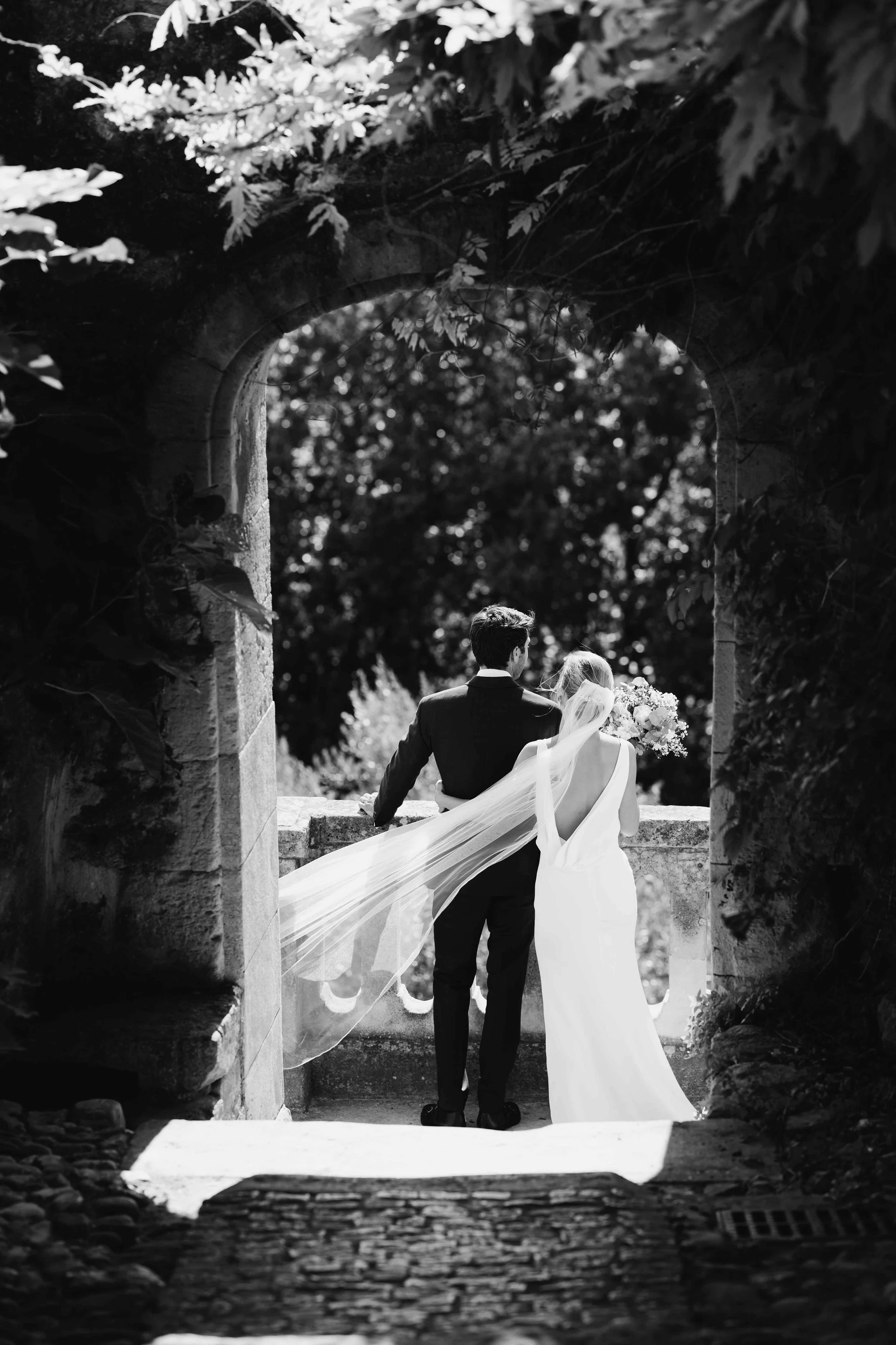 Un couple de mariés, vu de dos, dans le cadre d'une cérémonie nuptiale en plein air, encadrée par une arche en pierre et entourée de végétation, en noir et blanc. Domaine du bijoutier à grignan, drome provençal, coucher de soleil, session couple 