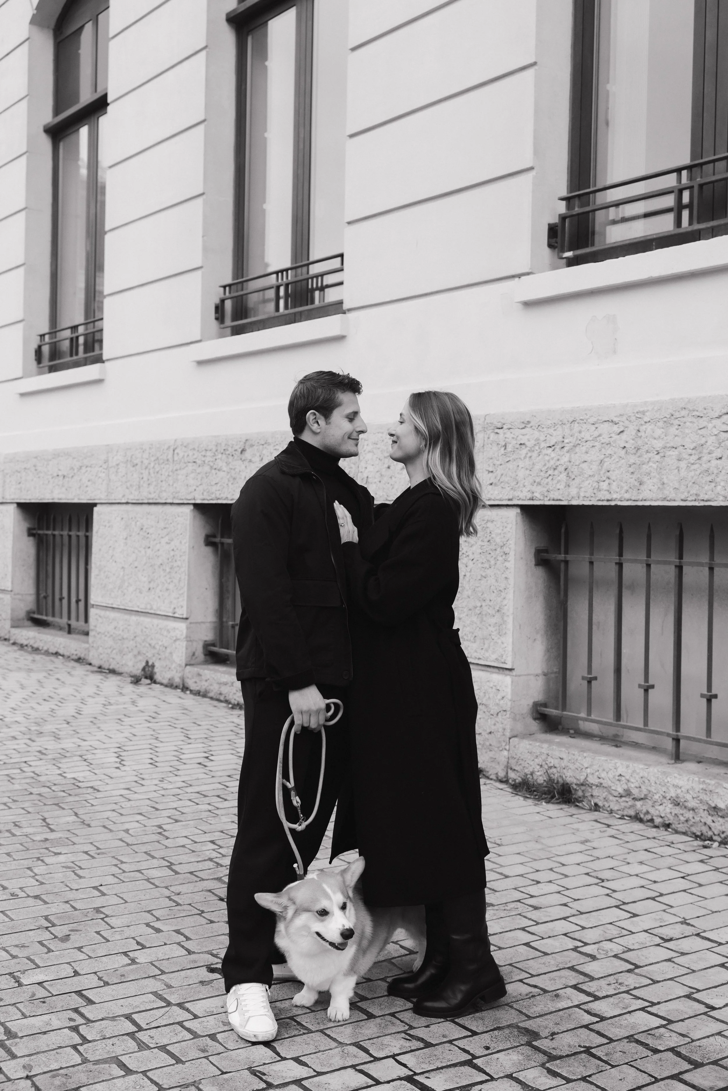 Un homme et une femme se regardent en souriant, debout sur un trottoir en pierre, avec un chien corgi entre eux. L'homme tient la laisse du chien. La scène est en noir et blanc, devant un bâtiment avec des fenêtres et des barres de sécurité. 