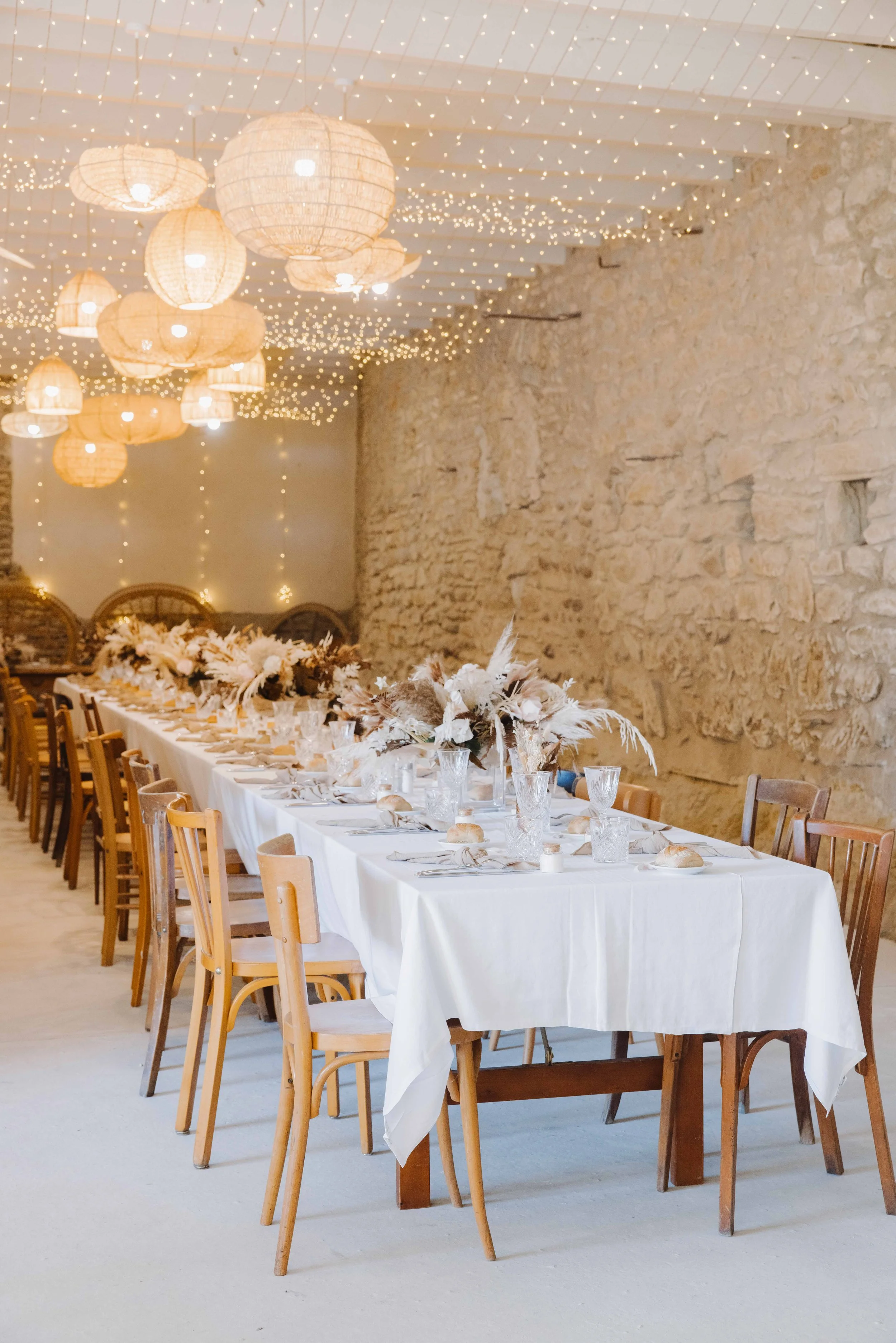 Salle de réception décorée pour un événement, avec une longue table élégante recouverte d'une nappe blanche, vaisselle transparente, bouquets floraux et des lampions en papier suspendus au plafond, ambiance chaleureuse et festive. Photographe mariage